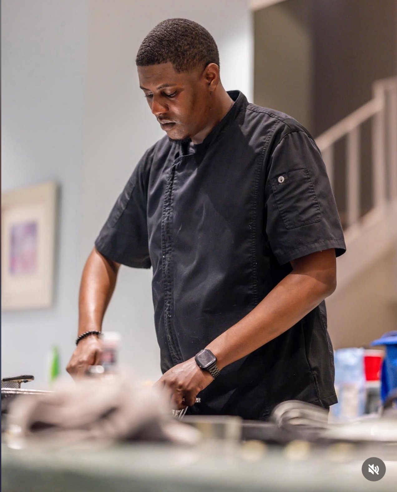 A man in a black chef's uniform appears to be preparing food in a kitchen or cooking area.