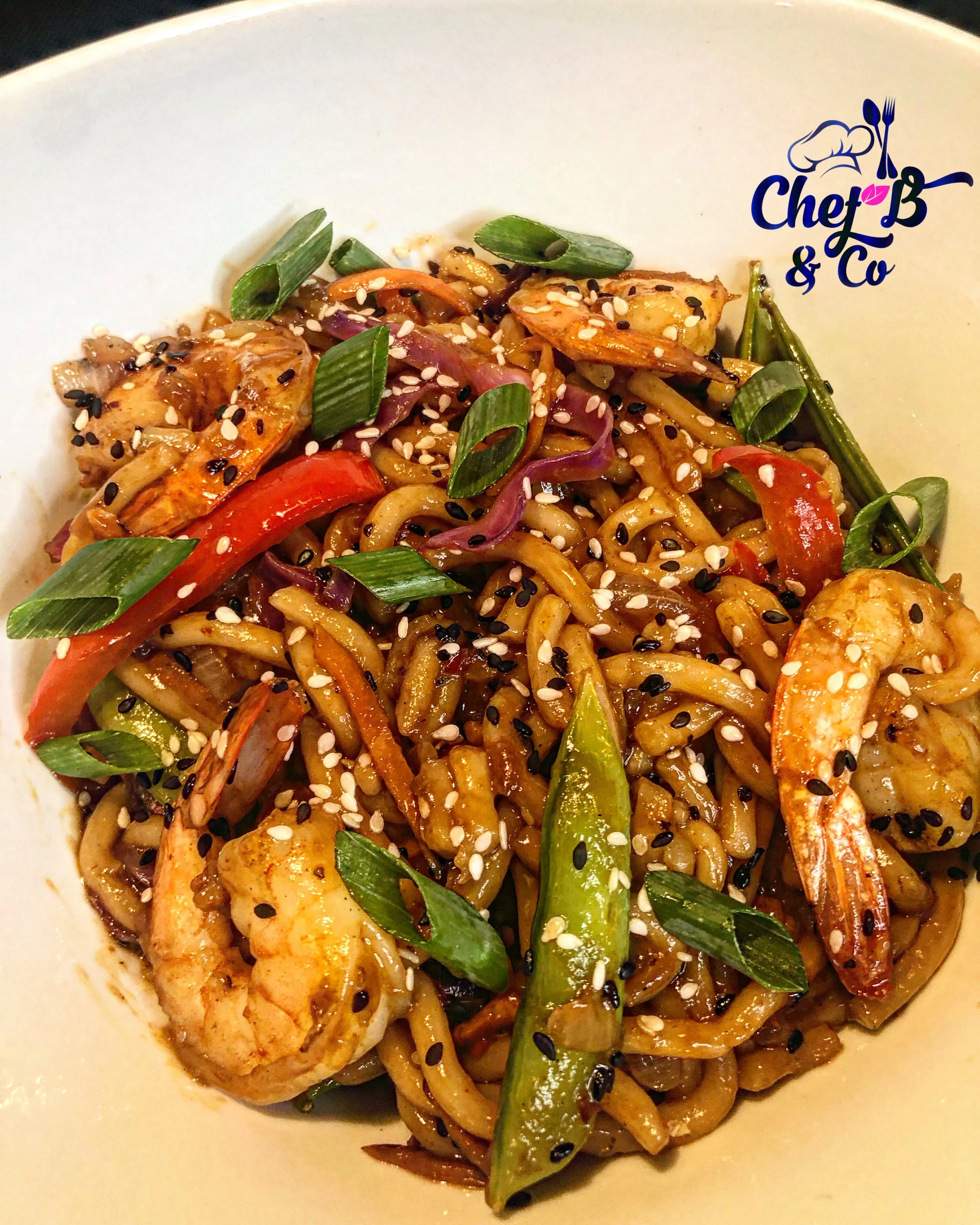 Close-up of a bowl of Asian-style stir-fried noodles with shrimp, red peppers, green onions, and sesame seeds topping.
