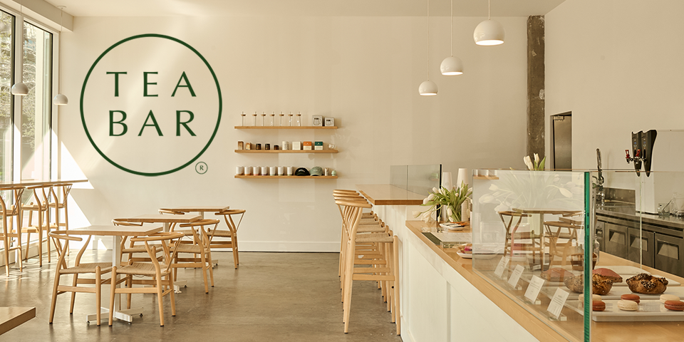 Interior of a teabar cafe with light wooden chairs and tables, a white counter with pastries, and a large green 'Tea Bar' logo on the wall.
