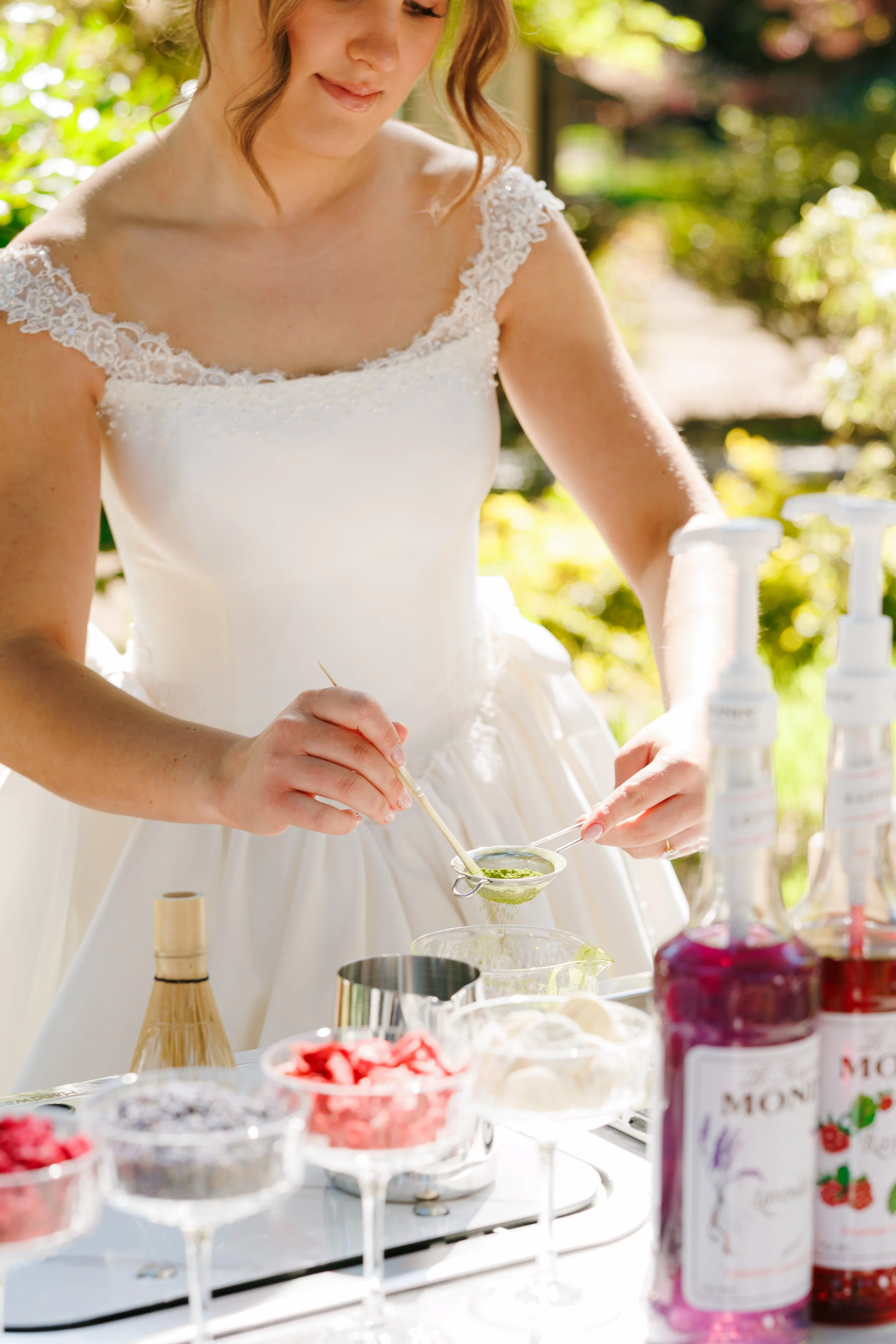 Woman in white dress preparing Matcha outdoors surrounded by jars and bowls with toppings and syrups.