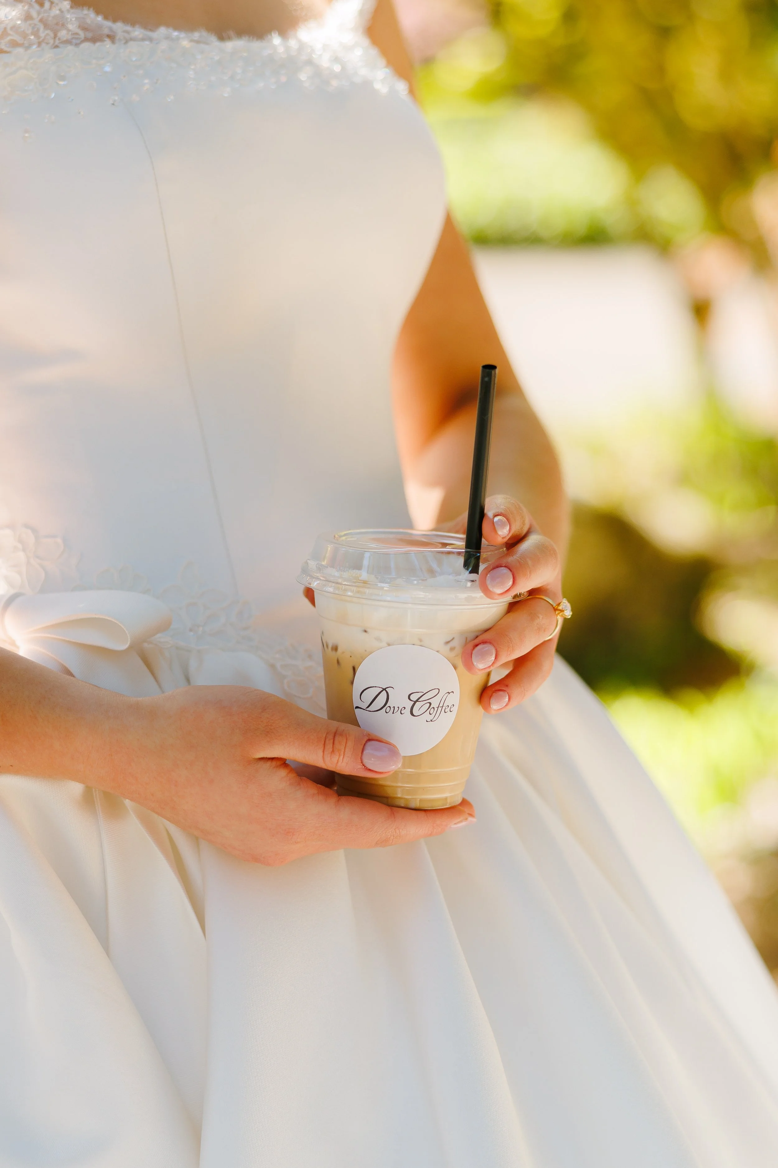 A person in a wedding dress holding a plastic cup of iced coffee with a straw and a Dove Coffee logo on the front of the cup.