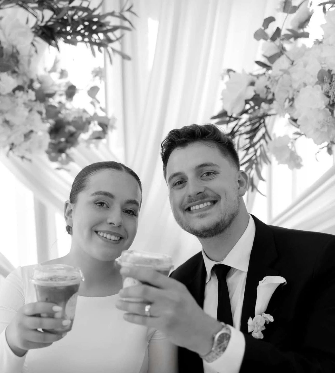 A happy couple dressed in wedding attire smiling and holding drinks at a celebration, with floral decorations and curtains in the background.