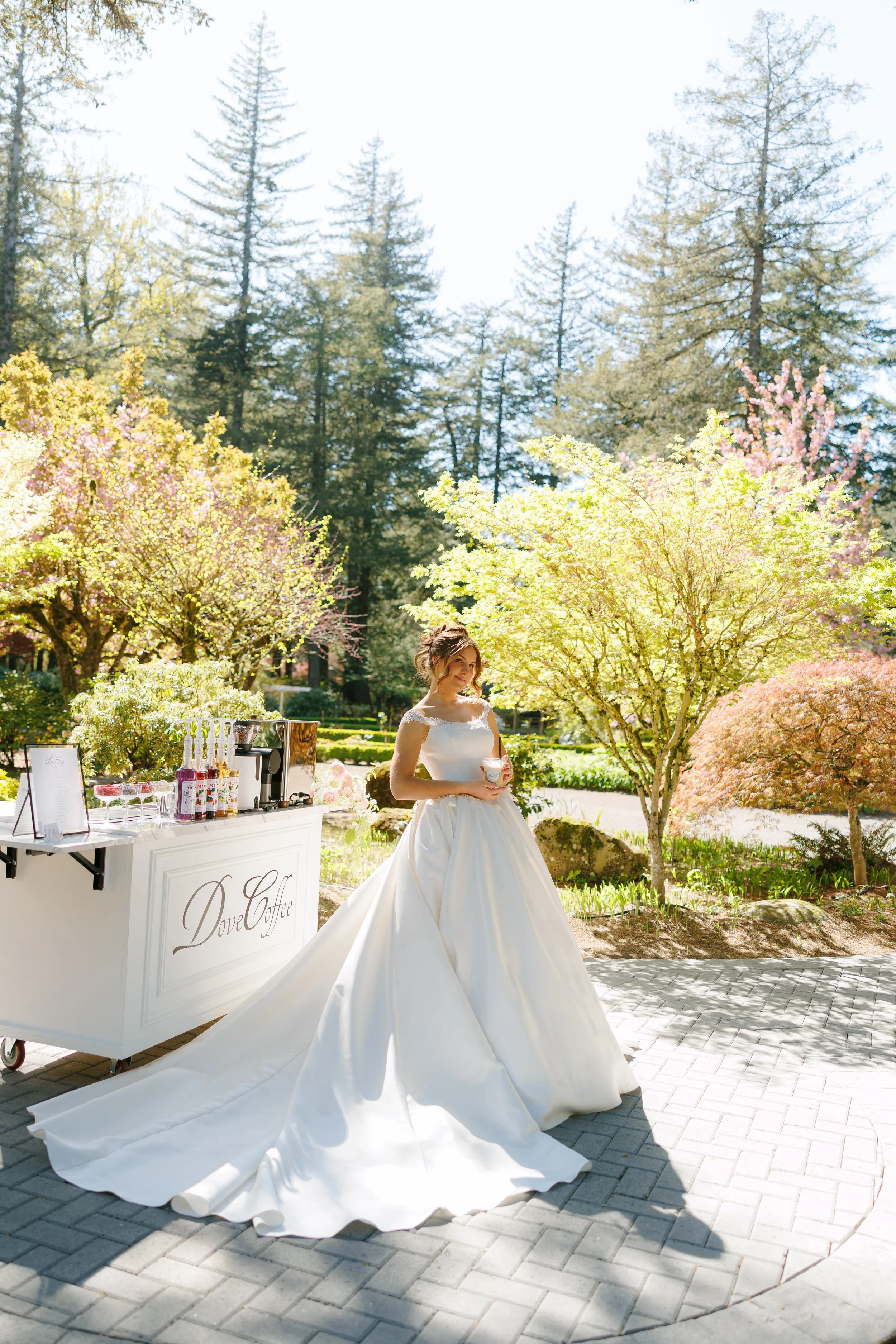 A bride in a white wedding gown holding a drink stands outdoors in a garden with trees and blooming flowers, next to a coffee stand labeled 'Dove Coffee' on a sunny day.