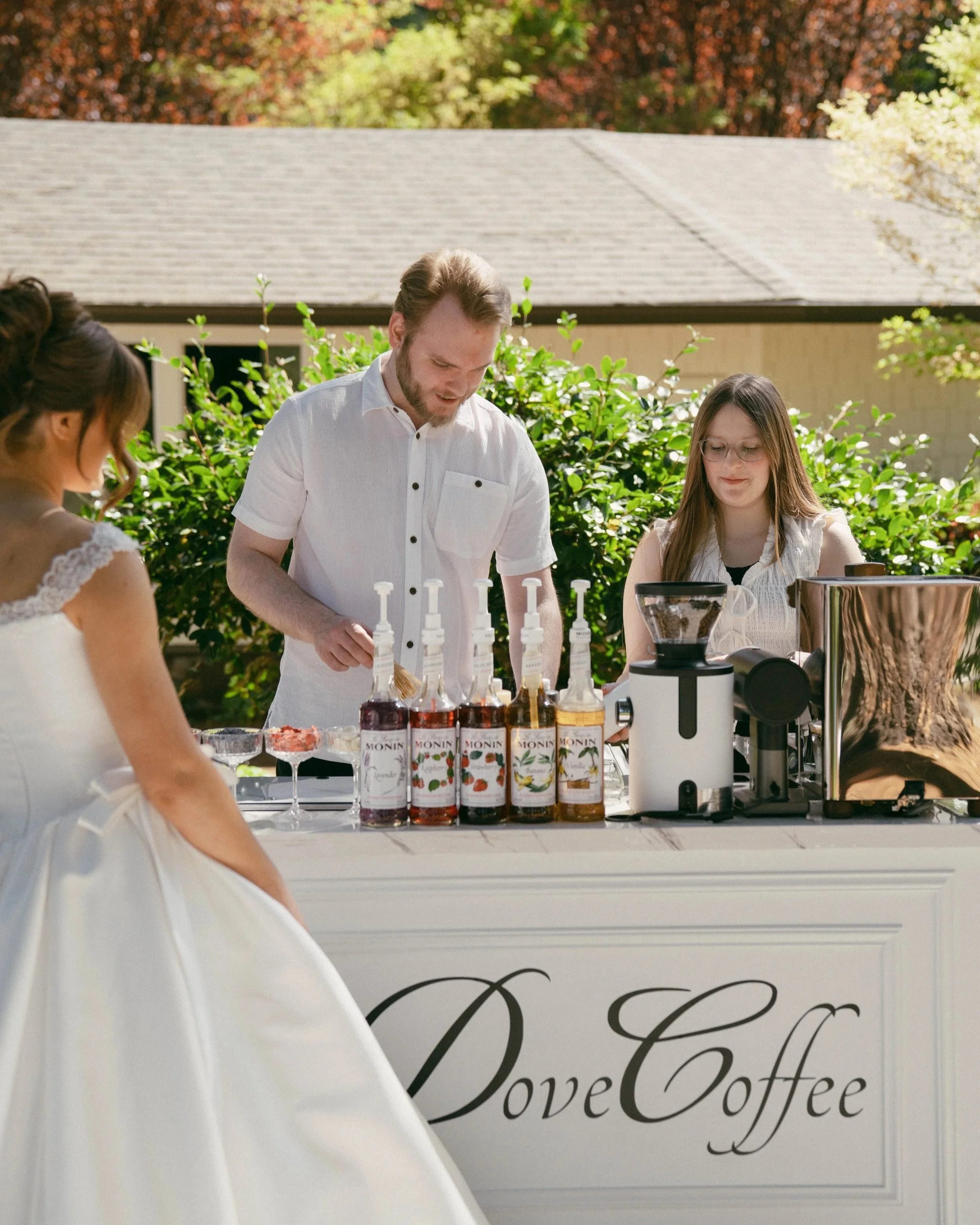 Husband and Wife making Matcha behind their coffee cart, 'Dove Coffee' as a bride in a white dress waits for her drink. All three people are smiling. Husband wears white shirt. Wife wears white laced shirt.