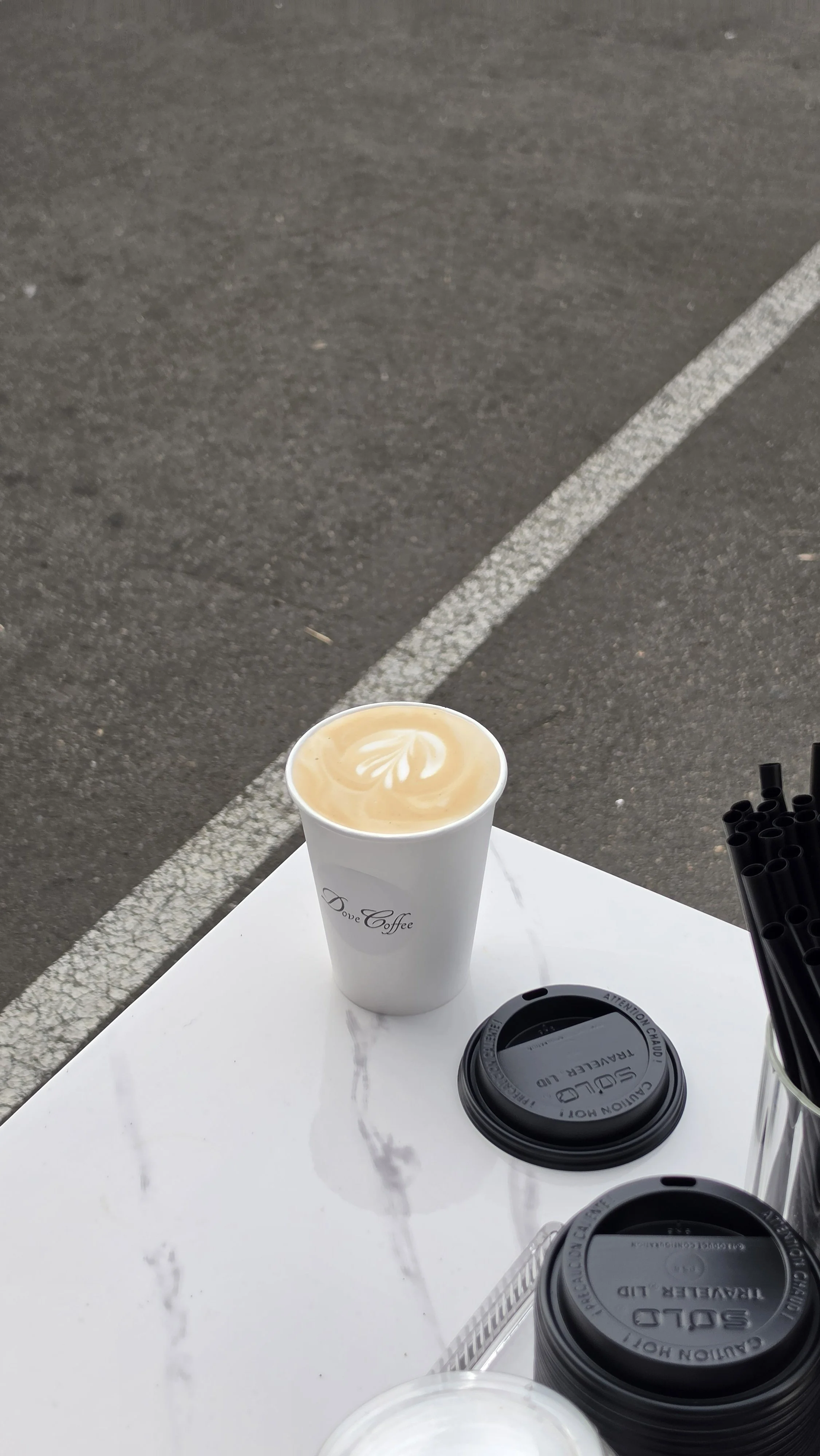 A to-go coffee cup with latte art on top, and a black plastic lid on a white table. There are other coffee cups and straws visible on the table.