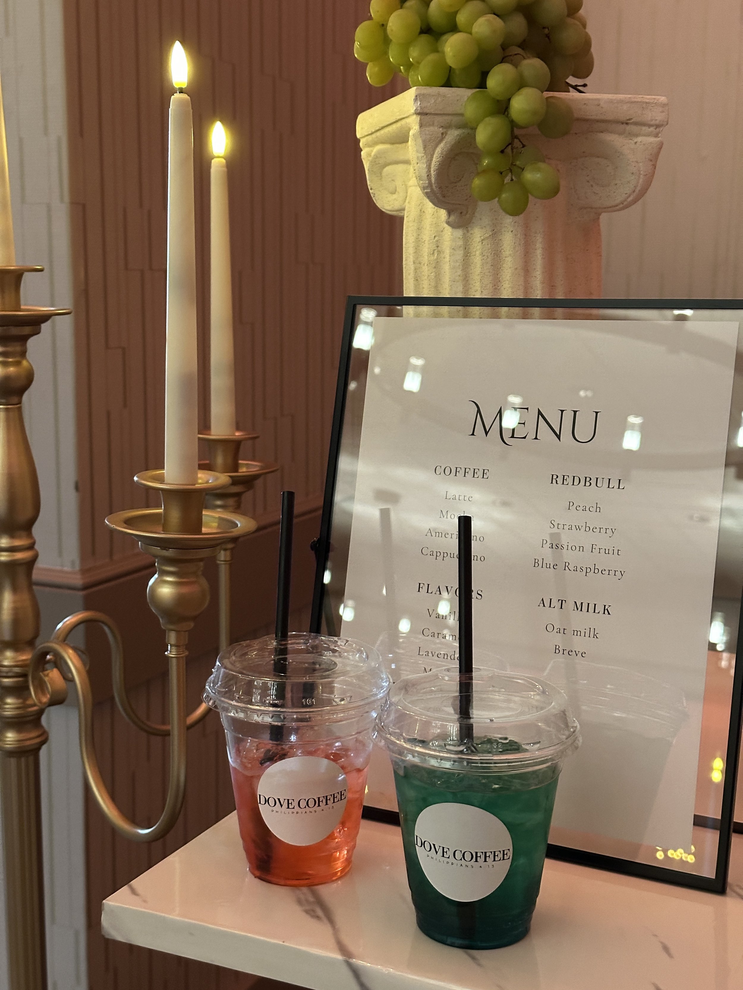 Two iced drinks in clear plastic cups with straws, one pink and one green, placed on a table beside a menu with coffee, redbull, and flavored options, in a cafe with candles and a decorative grapevine arrangement.