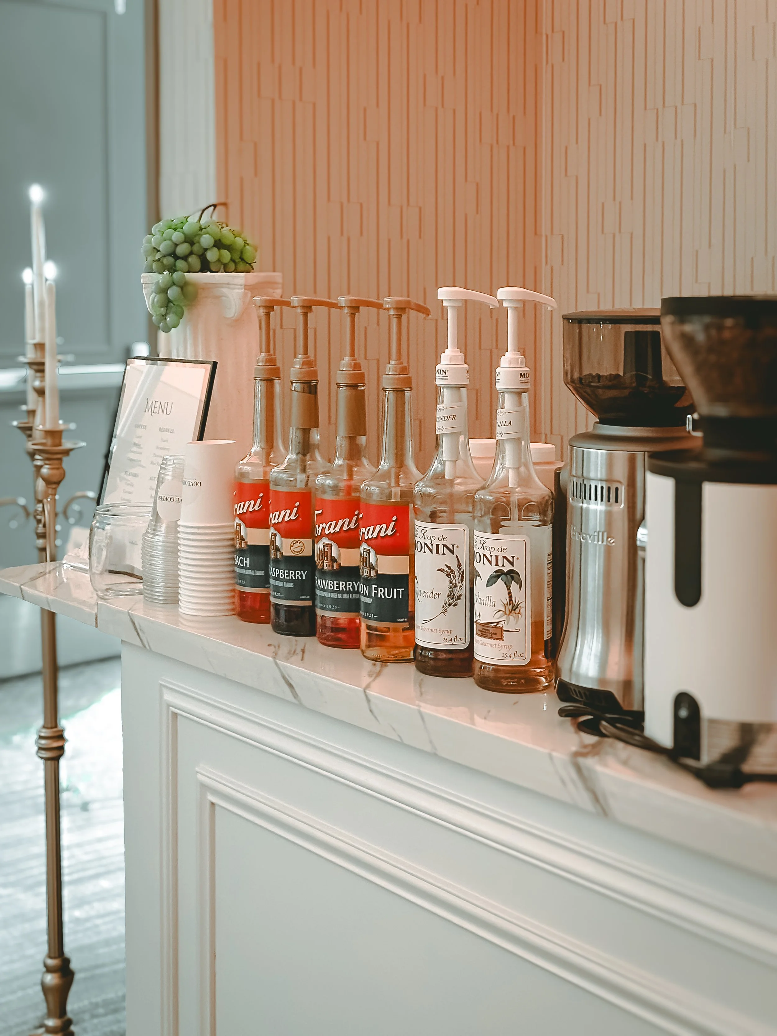 Assorted syrup bottles, paper cups, and a coffee machine on a counter in a cafe setting.