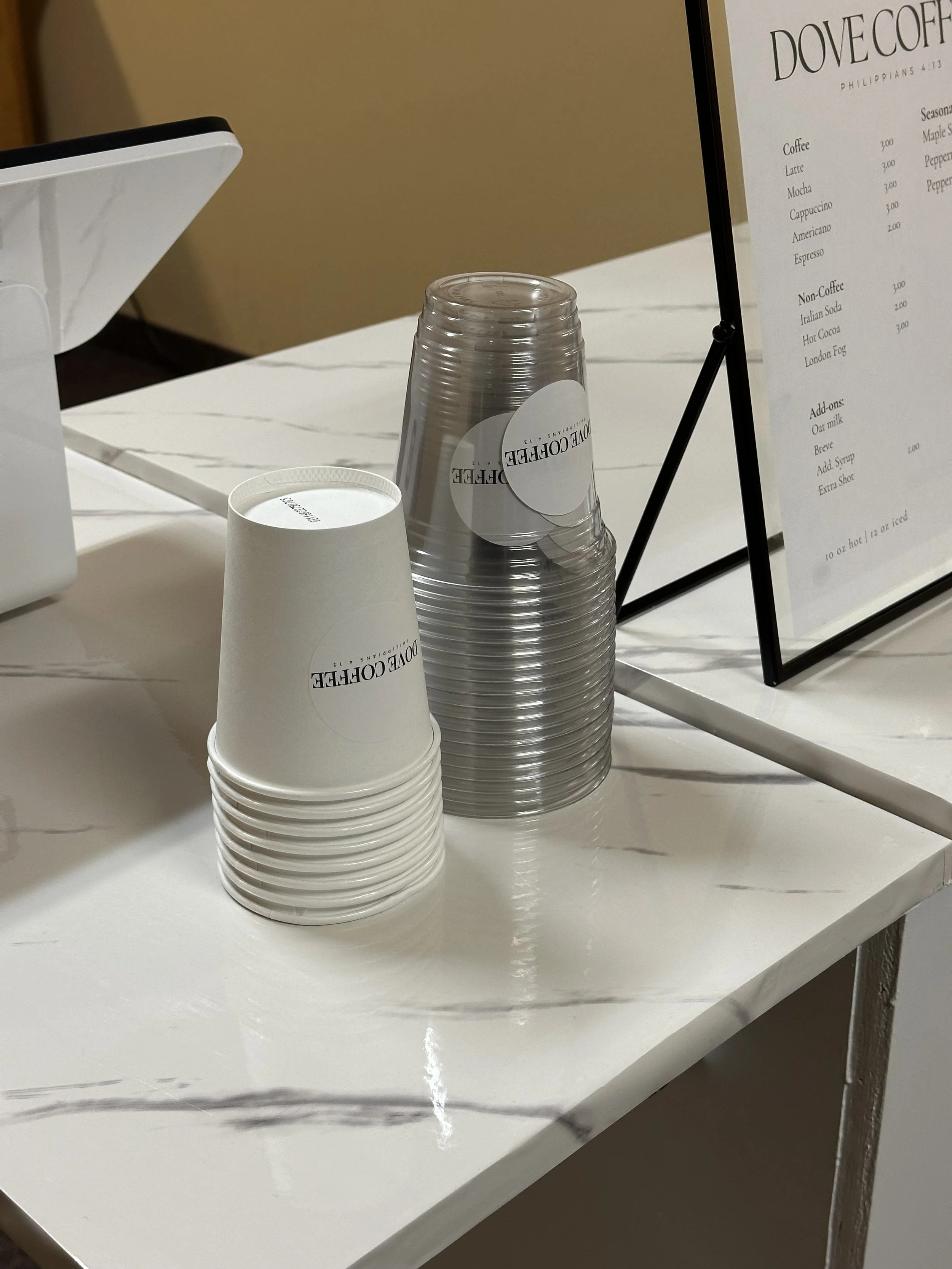 Stack of white paper cups and clear plastic cups on a white marble countertop at a coffee shop, with a black metal sign holder and a menu in the background.
