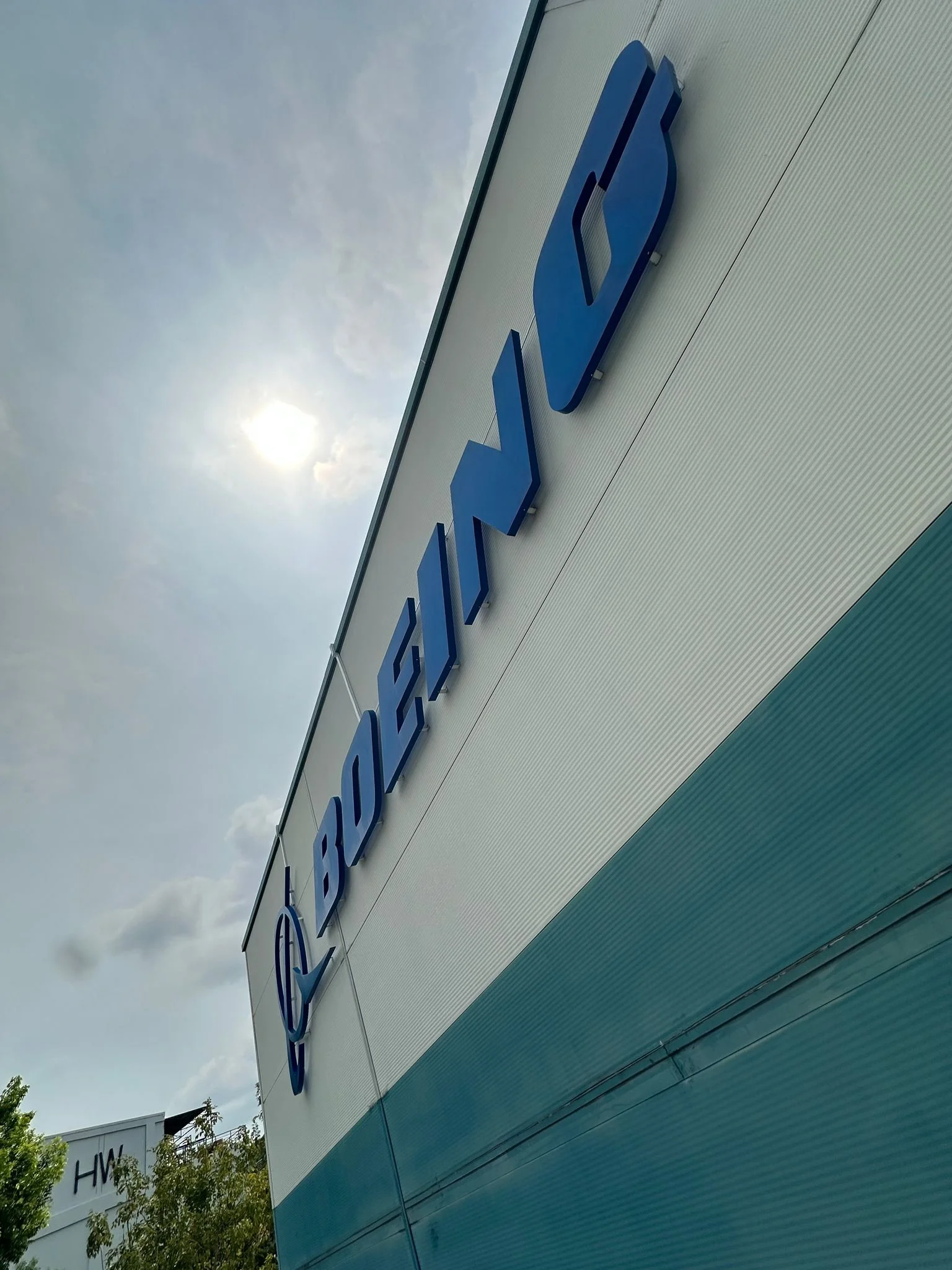 A large building with a sign that reads 'GE'. The building's exterior is white with a blue lower section. The sky is partly cloudy and the sun is visible behind clouds.
