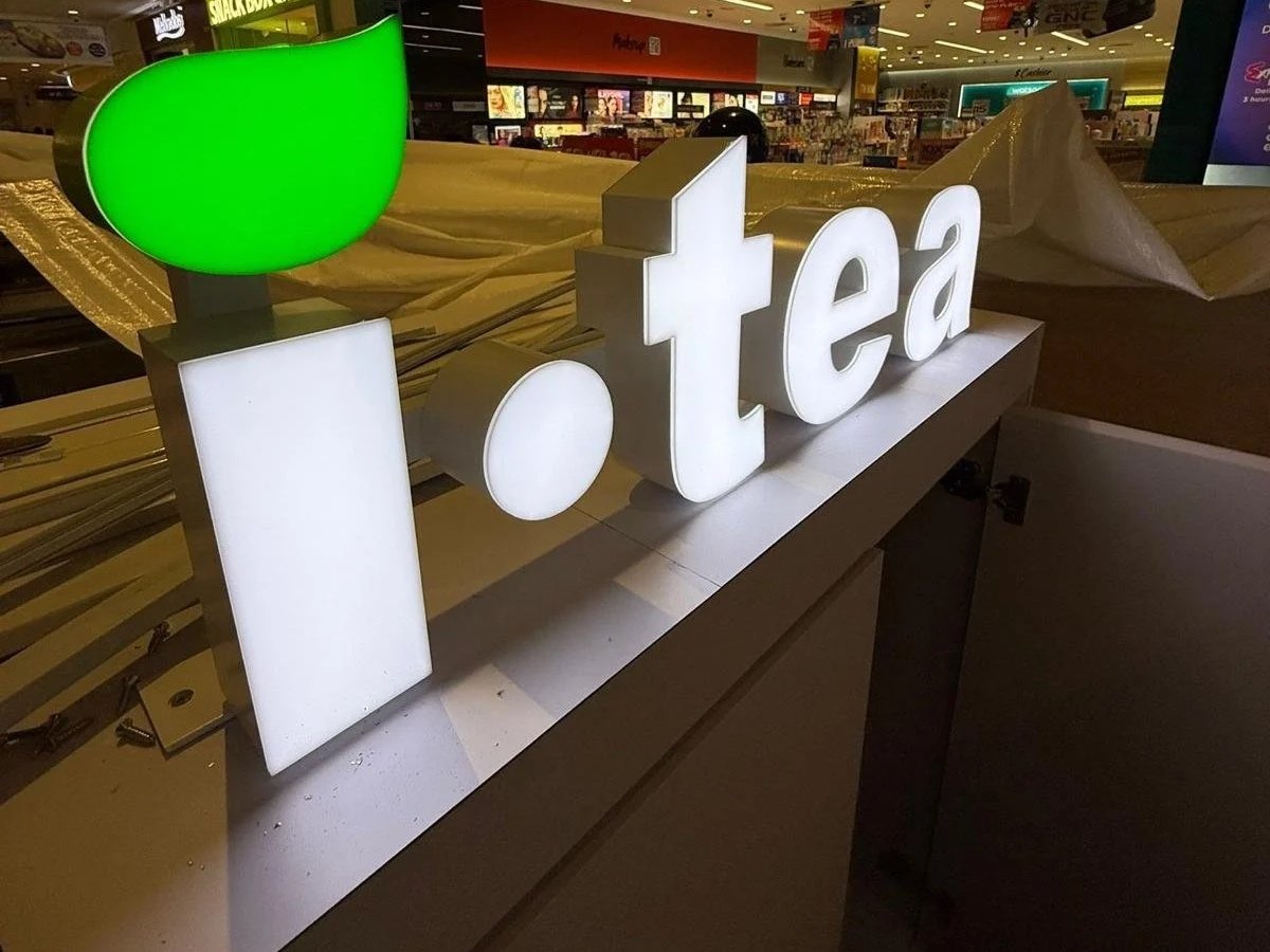 Illuminated white sign spelling 'tea' with a green leaf graphic, displayed on a counter in a shopping mall.