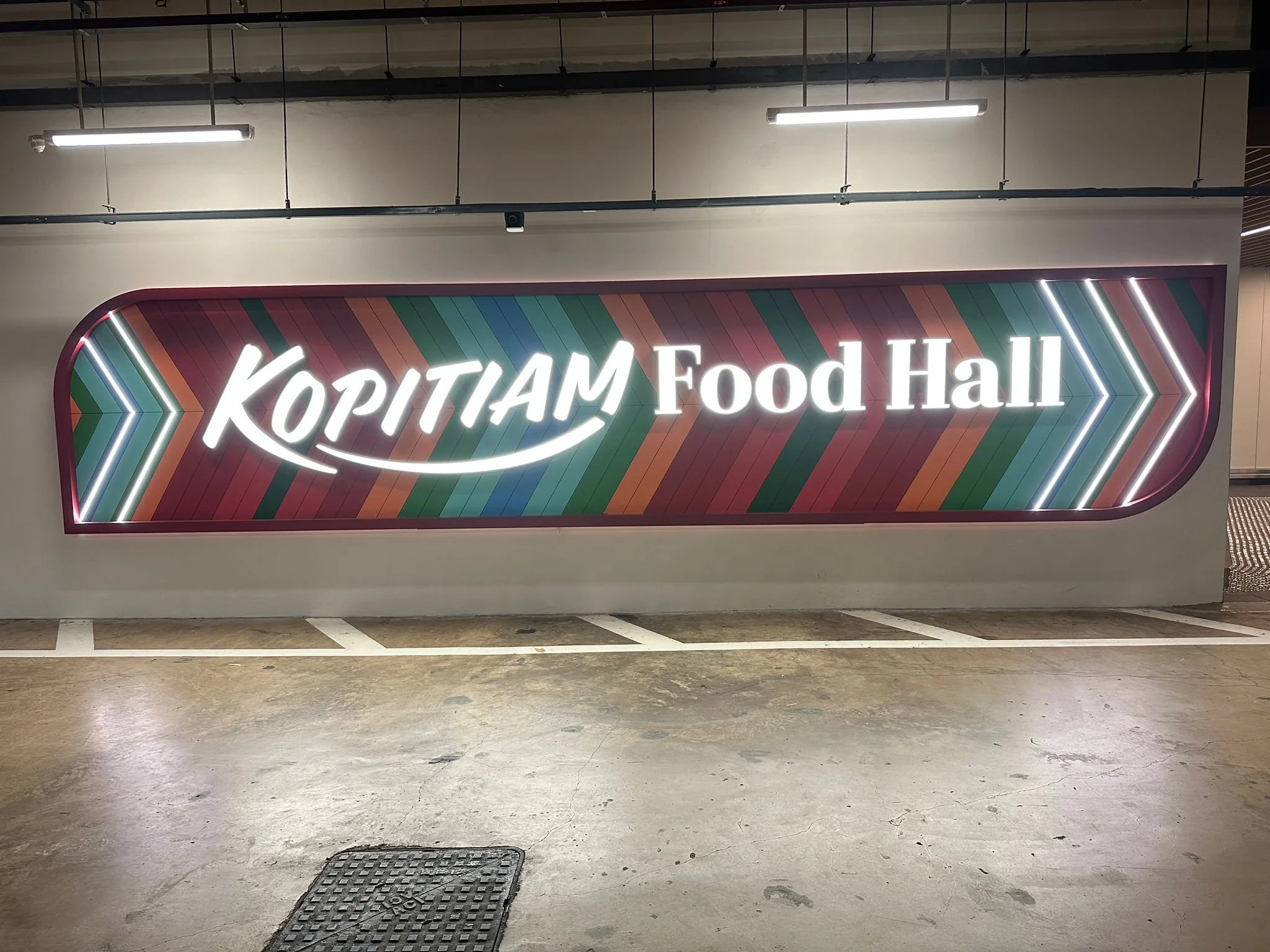 Sign for Kopitam Food Hall with colorful striped background and illuminated white text, located in an indoor parking garage.