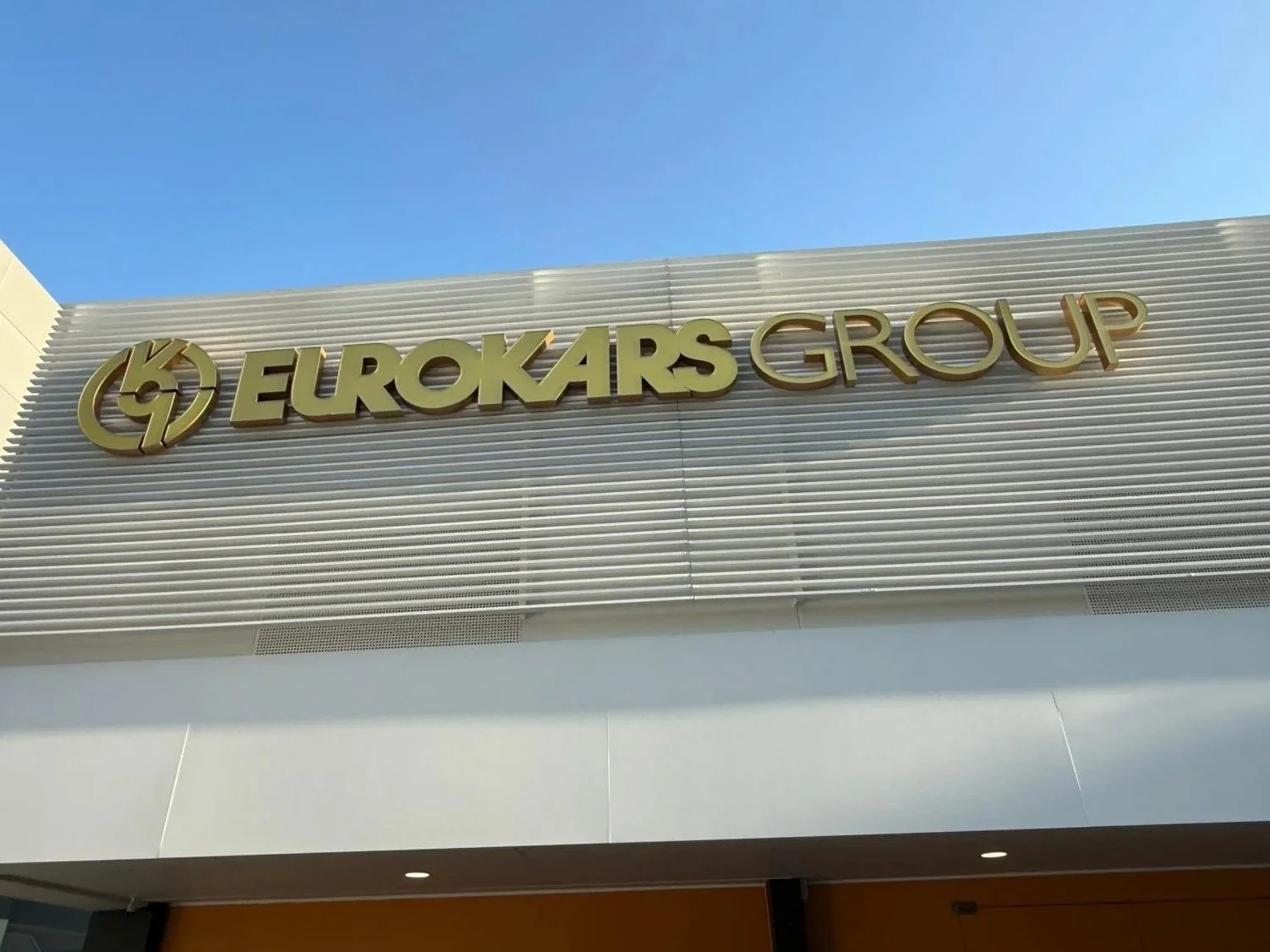 Exterior sign reading 'EUROKARS GROUP' with a logo featuring a lightning bolt inside a circular design, on a building facade under a clear blue sky.