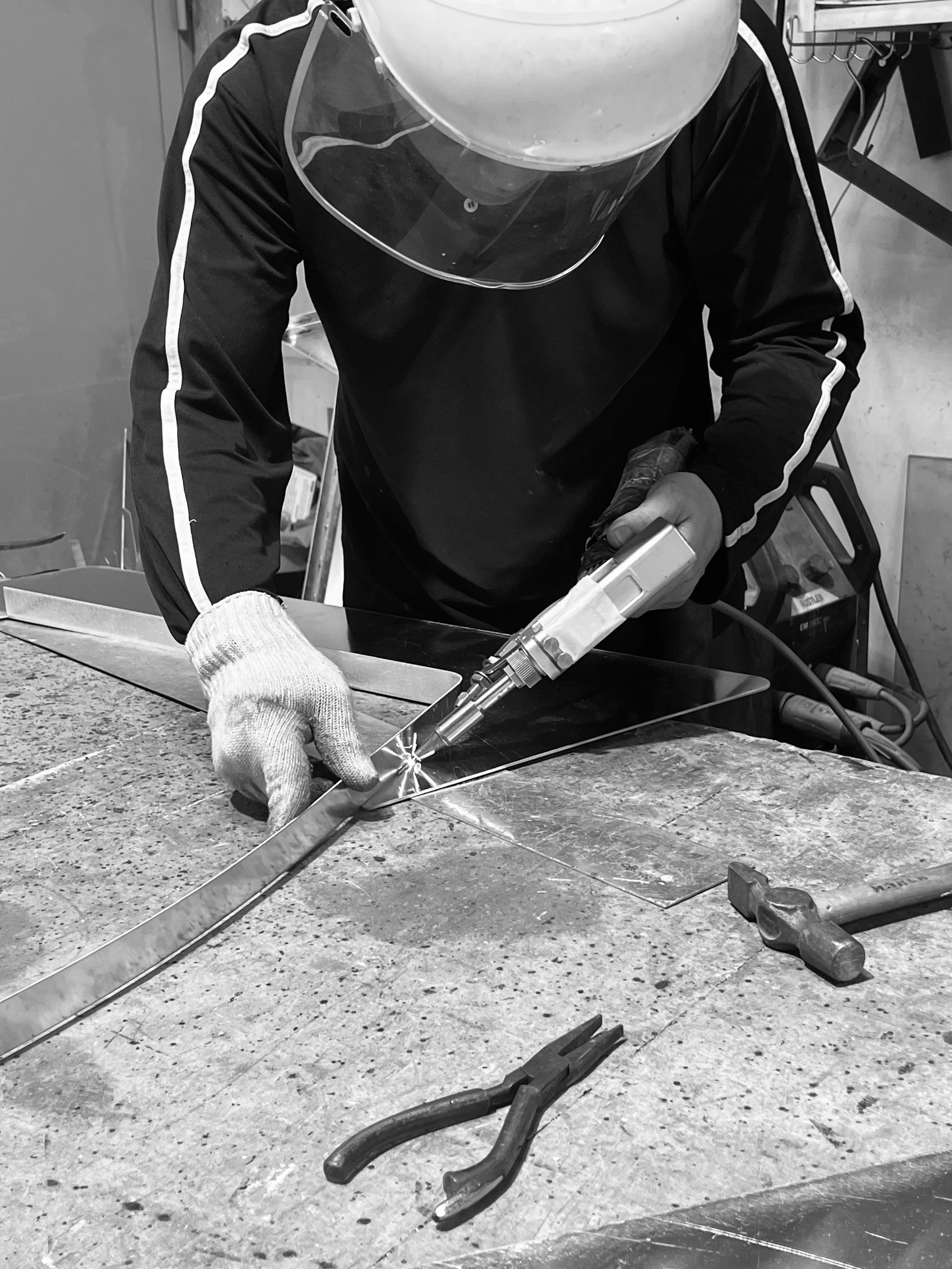 A person in protective gear welding metal on a worktable. The individual is wearing a welding helmet, gloves, and a safety jacket. The scene appears to be in a workshop or fabrication space with tools and equipment around.