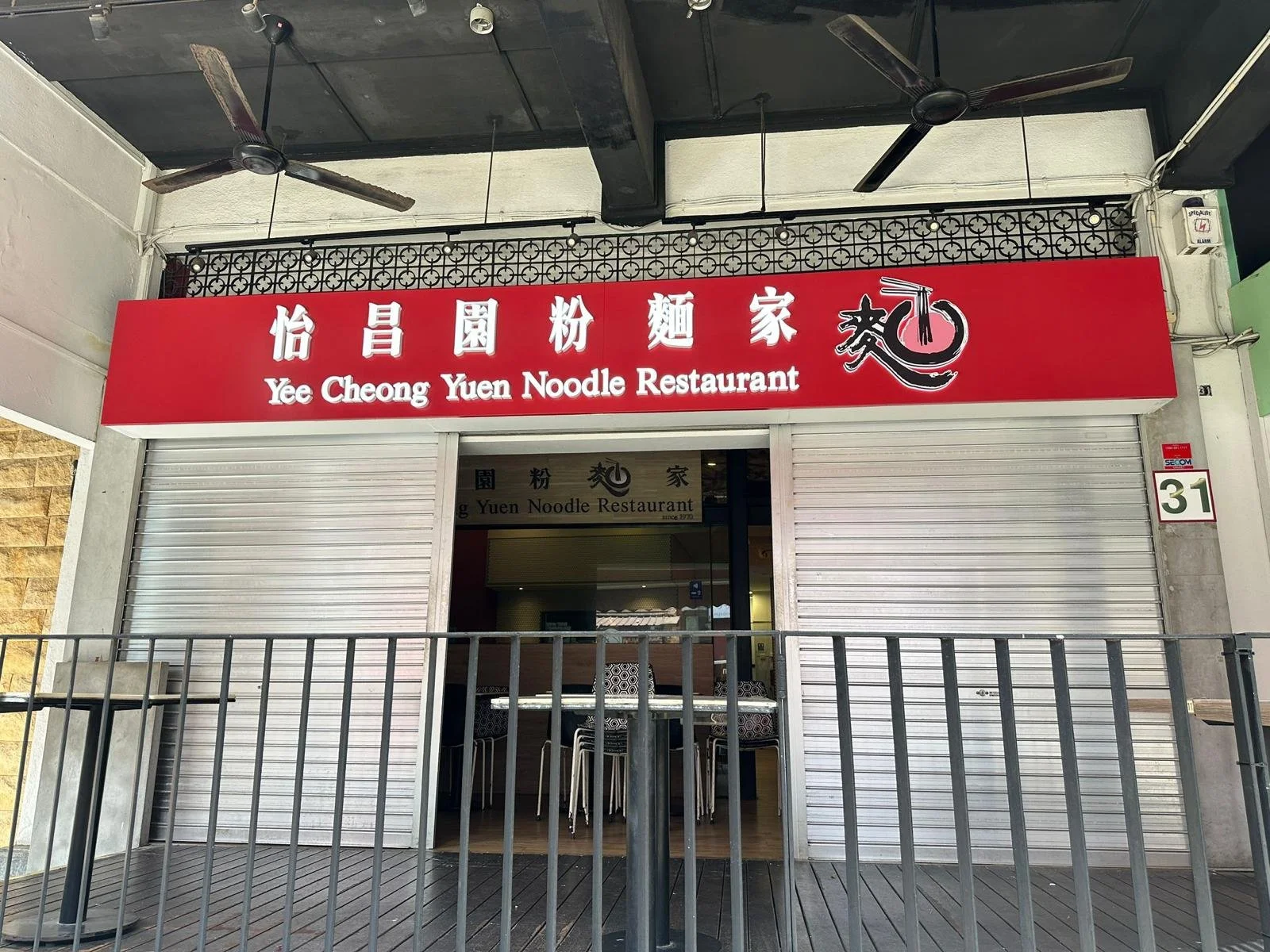 Closed restaurant with a red sign that has Chinese characters and the English name 'Yee Cheong Yuen Noodle Restaurant'. The restaurant has metal shutters, and there is a glass door with some chairs visible inside. A metal railing surrounds the entrance. The address number 31 is on the side. Two ceiling fans are visible on the ceiling.