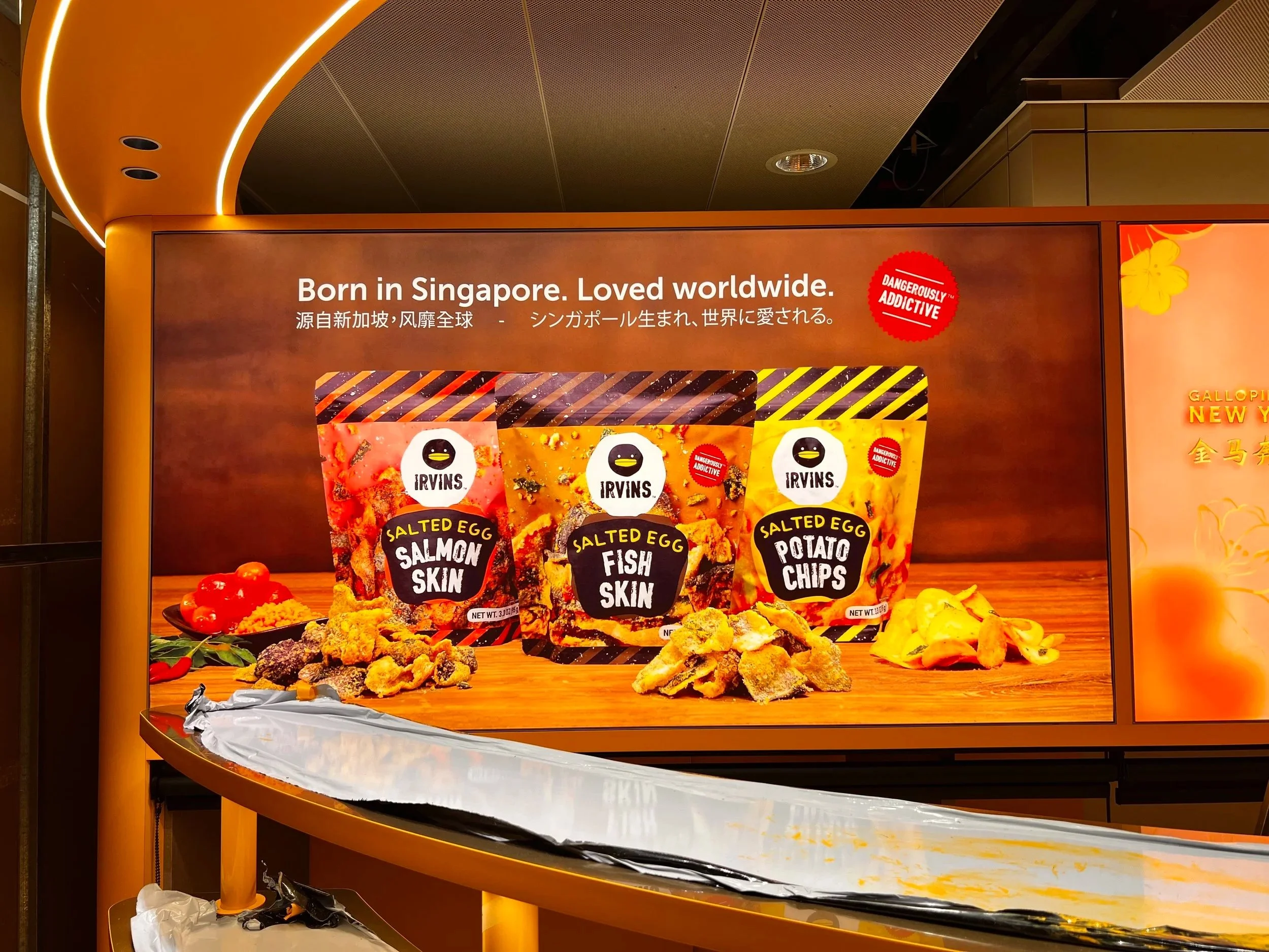 Display advertisement with the text "Born in Singapore. Loved worldwide" on top, featuring various snack products with labels "Salted Egg Salmon Skin," "Salted Egg Fish Skin," and "Salted Egg Potato Chips" along with a red sticker that says "Dangerously Addictive," and some snacks placed on a table with scattered packaging and decorations.