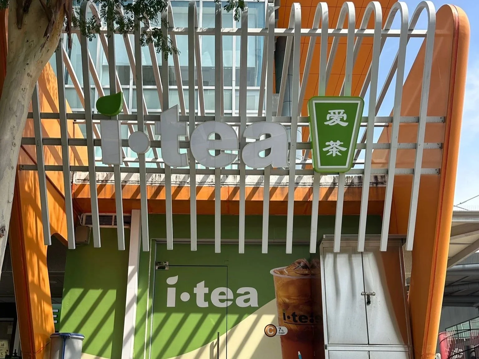 Signboard of a tea shop named 'i·tea' with a green and white color scheme, and a small green logo resembling a tea leaf, with a background of a modern building.