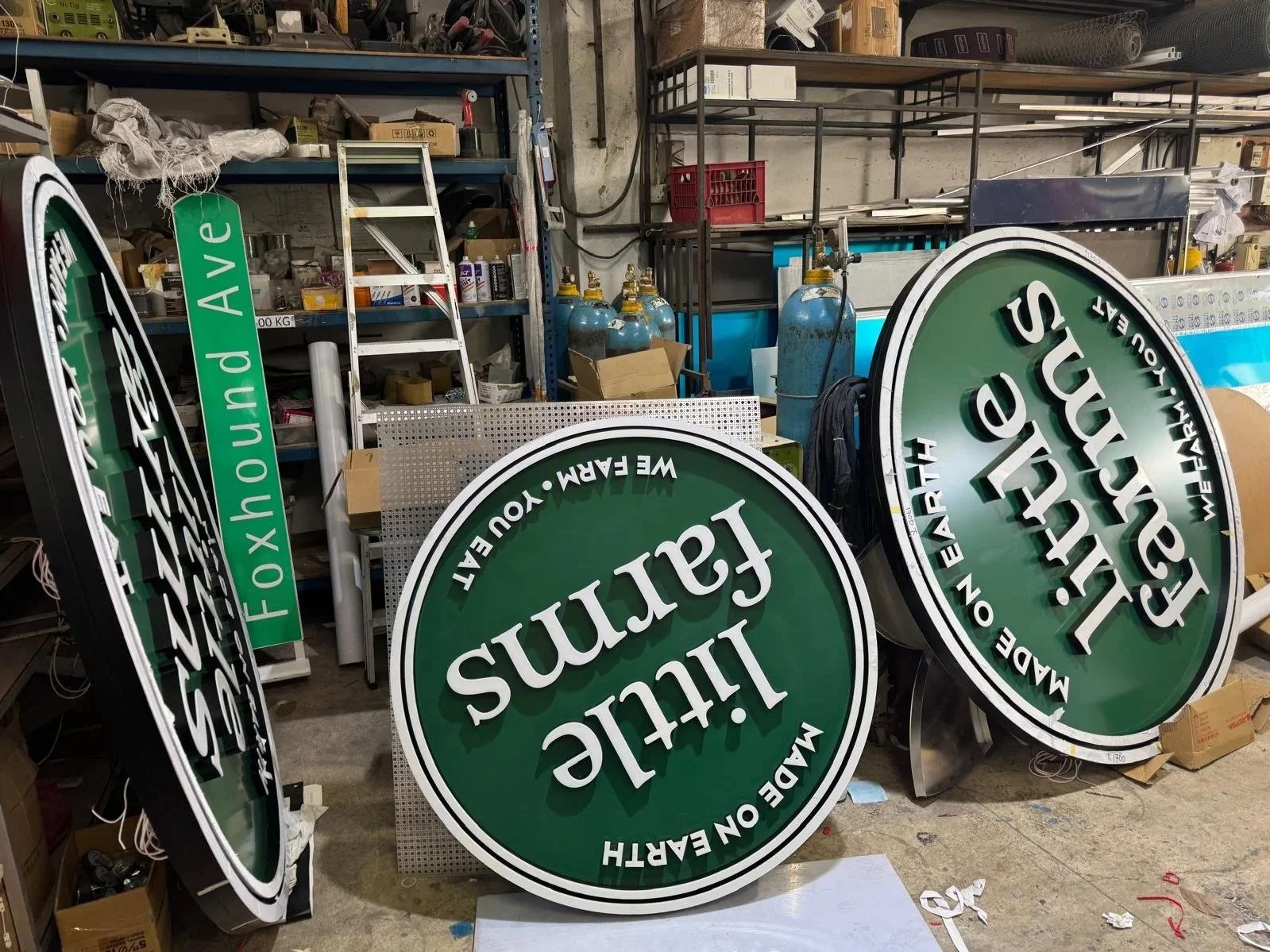 Workshop with multiple illuminated green and white signage that read "Lucille's Faucets" and "Made on Earth!" in a cluttered space with shelves, gas tanks, and various supplies.