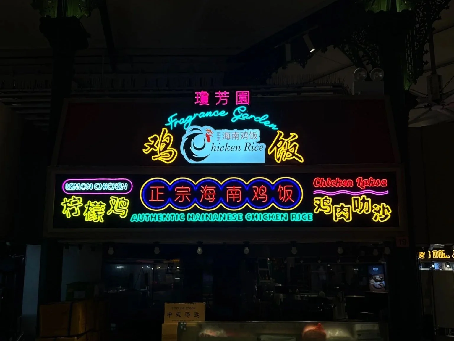 Neon sign advertising a chicken rice restaurant named Fragrance Garden with Chinese characters, and phrases like 'Authentic Hainanese Chicken Rice', 'Chicken Laska', and 'Lemon Chicken' in various bright colors.