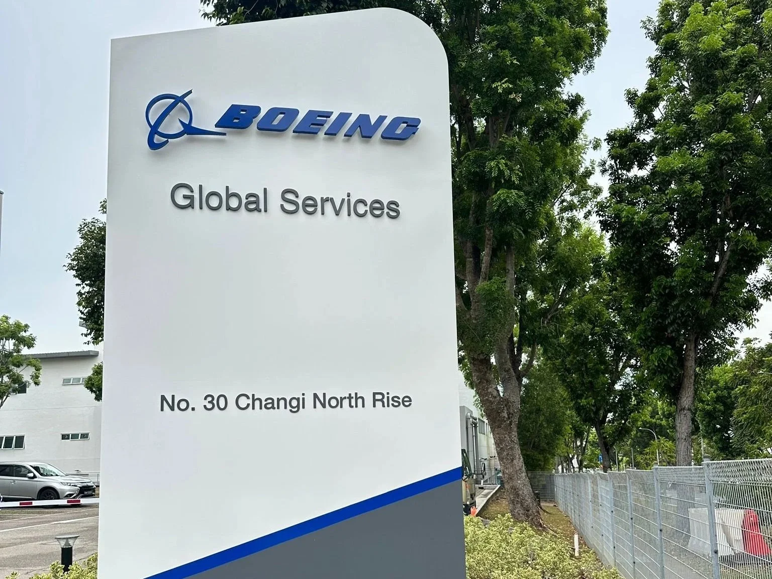 Signboard with Boeing logo and text, reading "Boeing Global Services, No. 30 Changi North Rise," situated outdoors among trees and a parking lot.