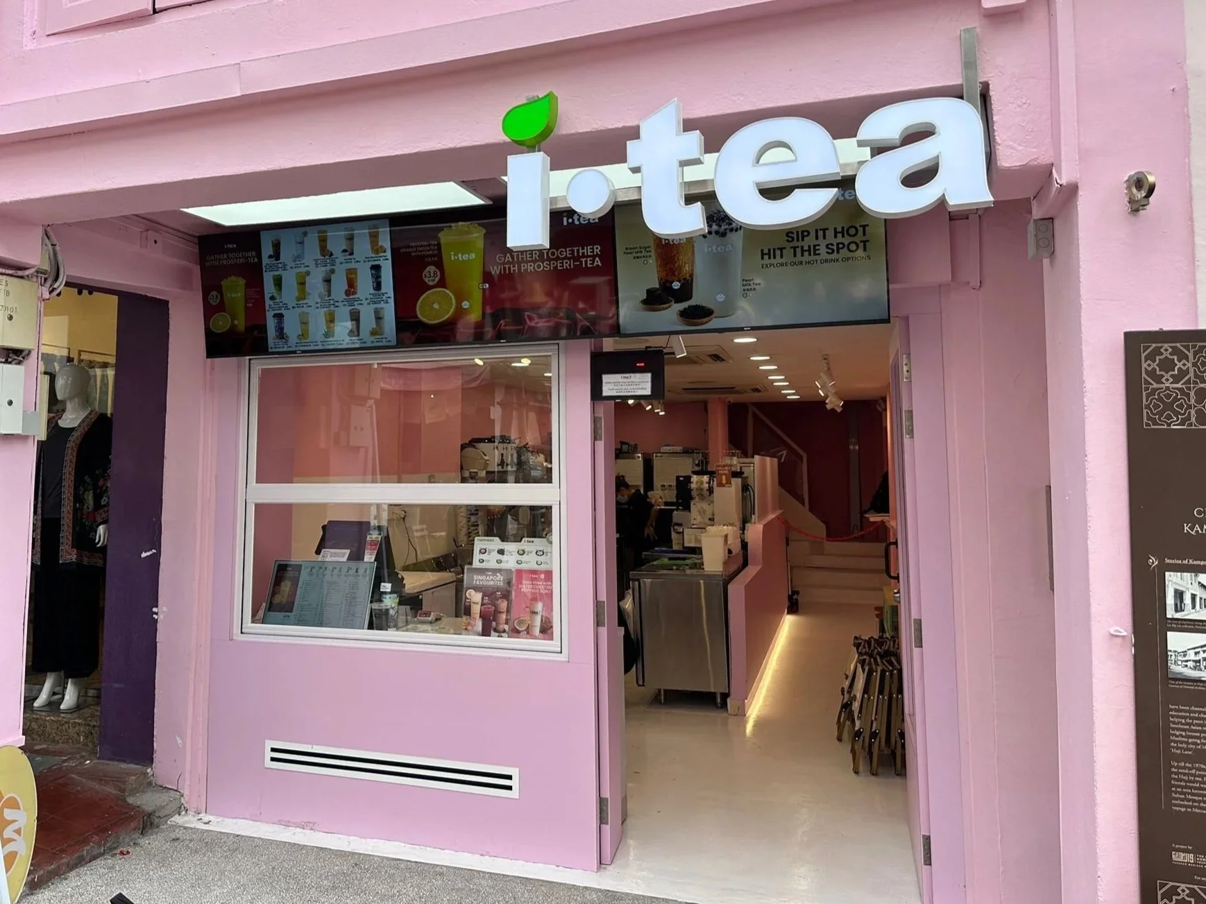 Front of a pink tea shop with a white sign that reads 'i-tea' and green leaf logo, open entrance showing interior with customers.