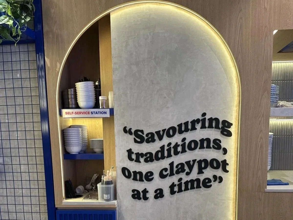 A sign with quote 'Savouring traditions, one claypot at a time' next to a self-service station with bowls, utensils, and condiments in a restaurant.