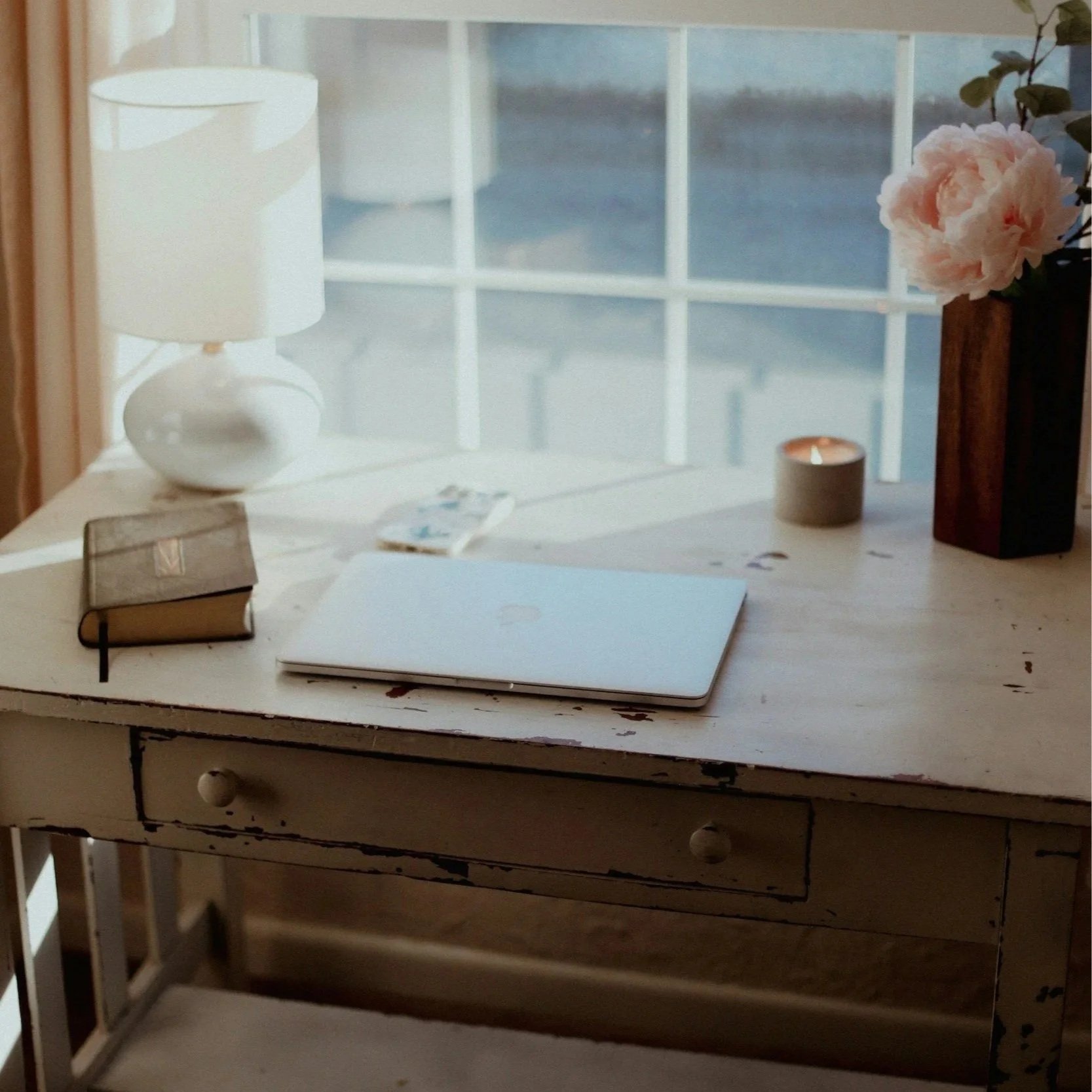 A rustic white wooden desk with a laptop, closed, on top. To the left of the laptop is a small notebook with a black bookmark. Behind the notebook is a white table lamp with a round base. To the right of the laptop, there is a small candle and a black vase with a pink peony flower. In the background, there is a window showing the outdoor scene with grass and a fence.