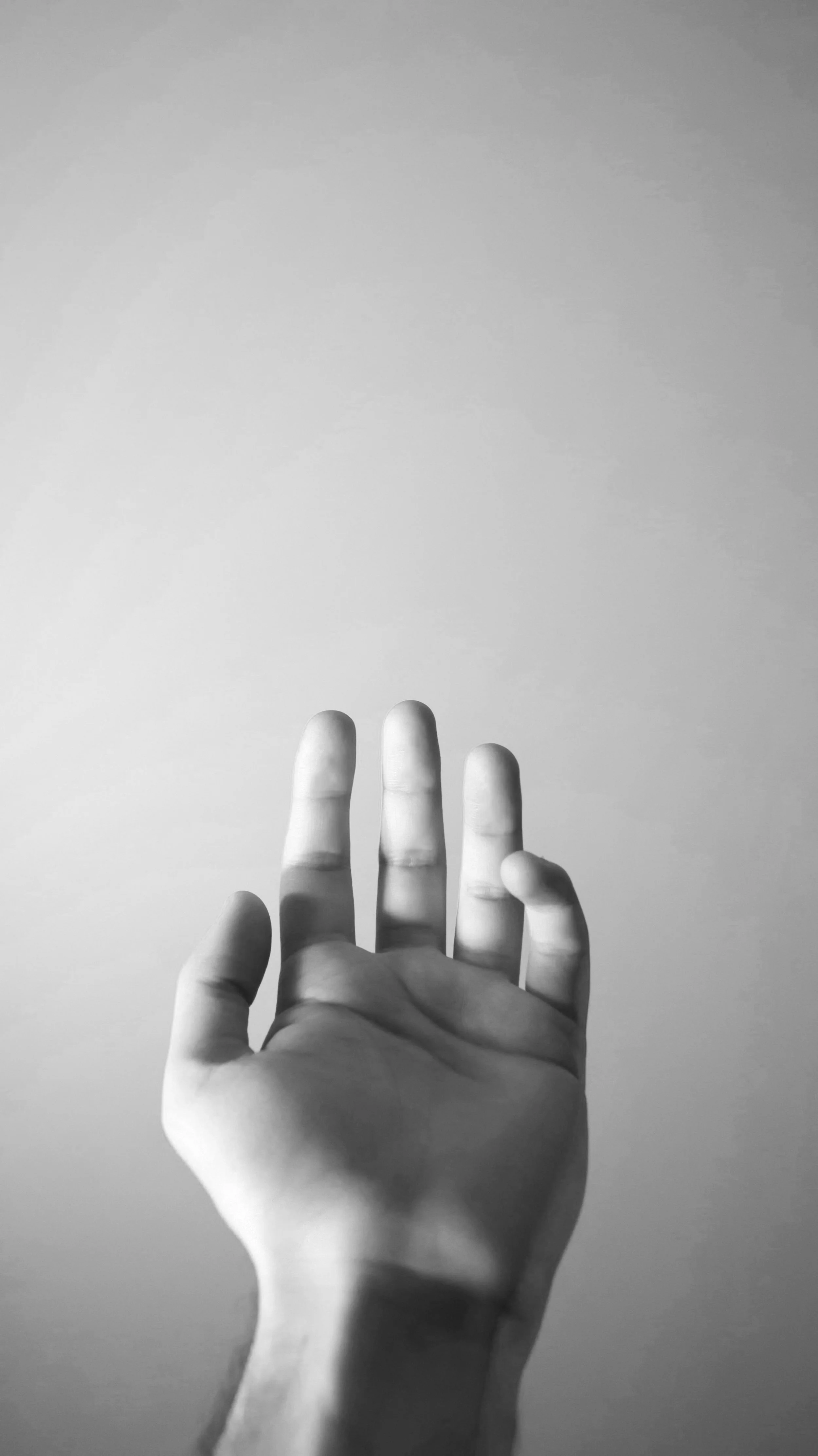 Black and white photograph of a person's hand with fingers slightly spread, reaching towards a plain, light-colored background.