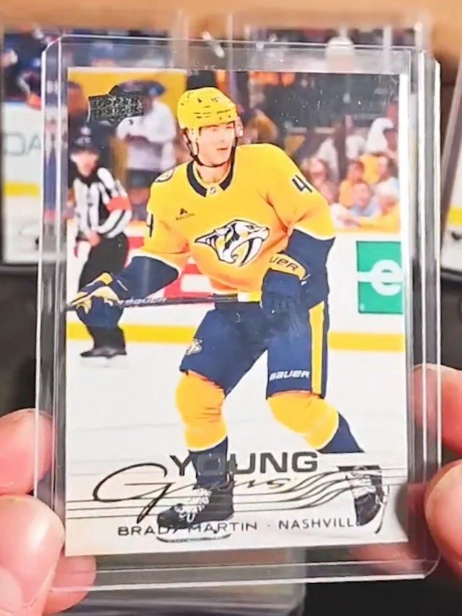 A trading card featuring a young hockey player in a yellow Nashville Predators uniform, holding a hockey stick during a game, with an arena background and a crowd in the stands.