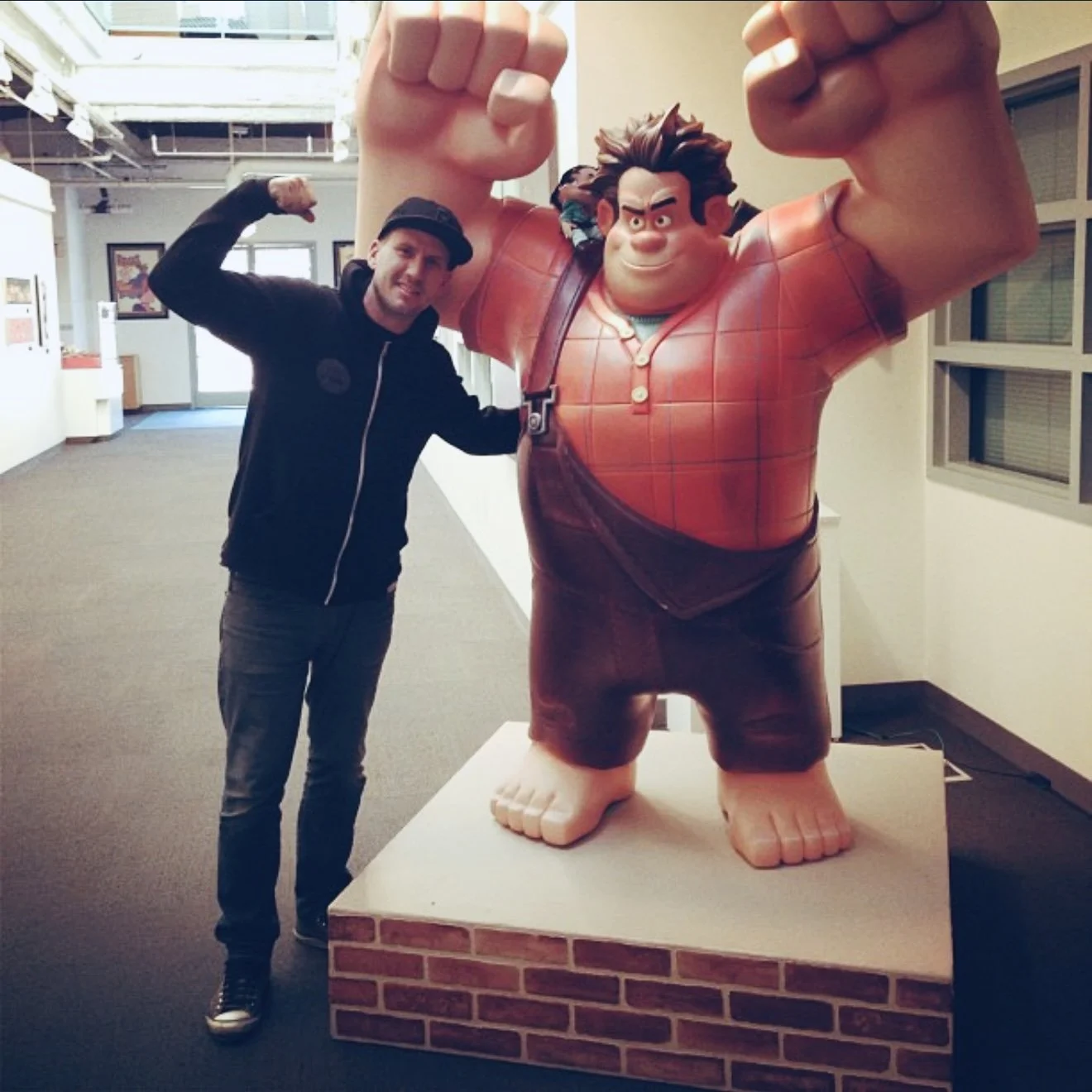 A man in casual clothing and a black hat standing next to a large cartoon statue of a muscular man with glasses, wearing a red plaid shirt and brown shorts, raising a fist.