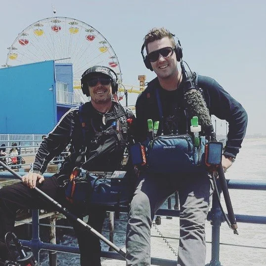 Two men wearing headsets and headphones smiling at the camera, standing on a pier with an amusement park and a Ferris wheel in the background.