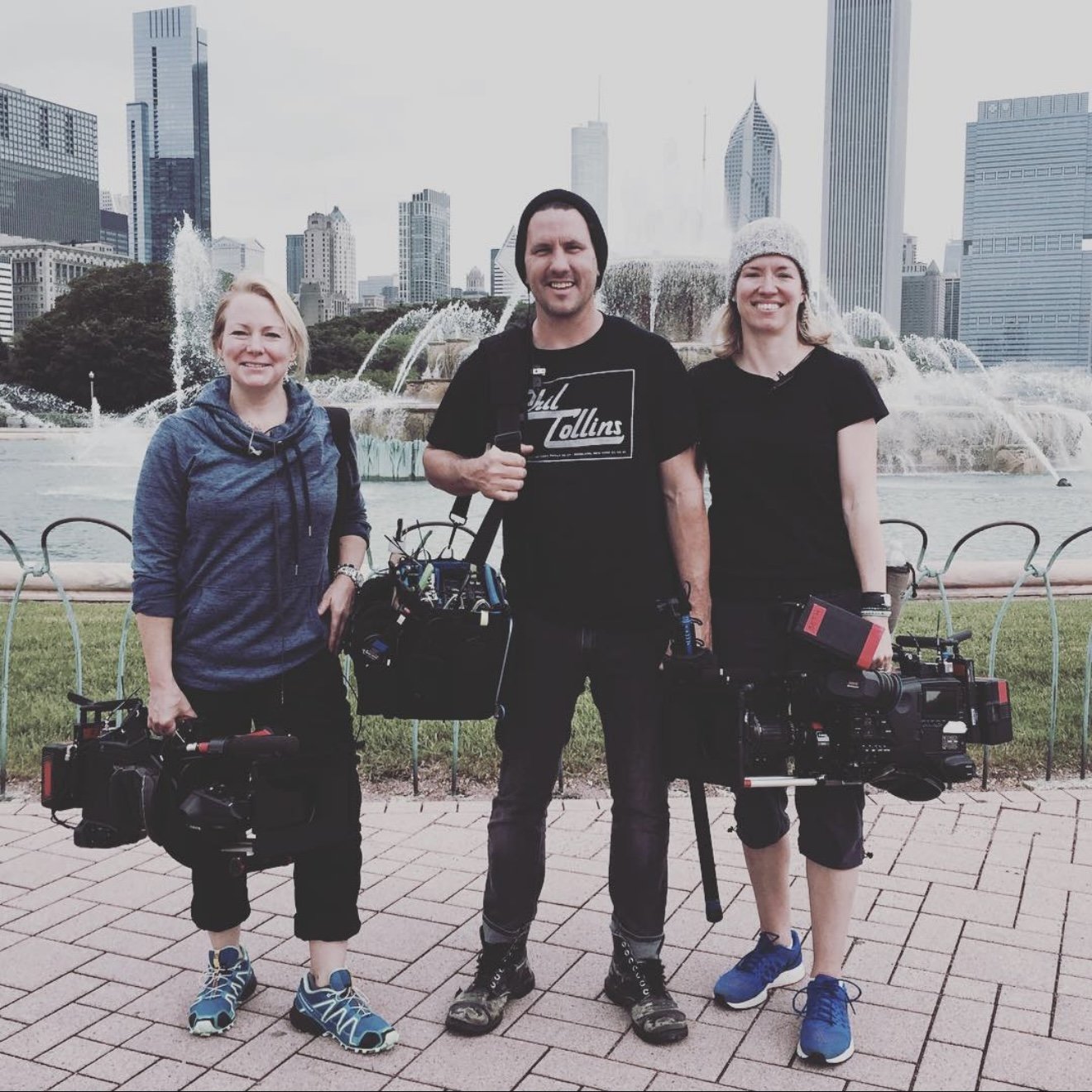 Three smiling cameramen standing outdoors in front of a city skyline with a fountain, holding professional camera equipment.