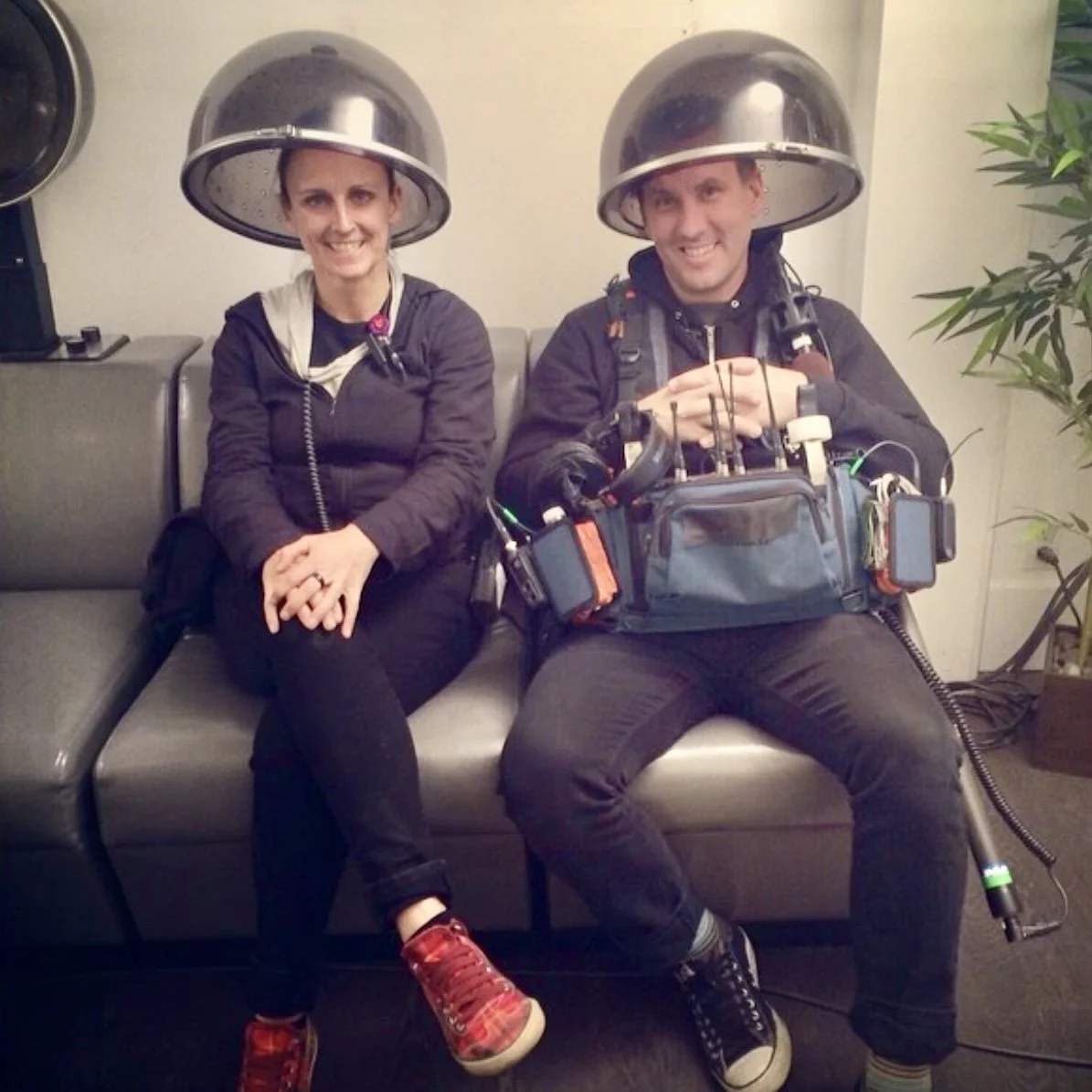 A man and woman sitting on a bench with hair salon hood dryers over their heads, smiling at the camera. The woman is on the left, wearing a black jacket and red sneakers, with her hands clasped. The man on the right has a rehab or medical device kit 