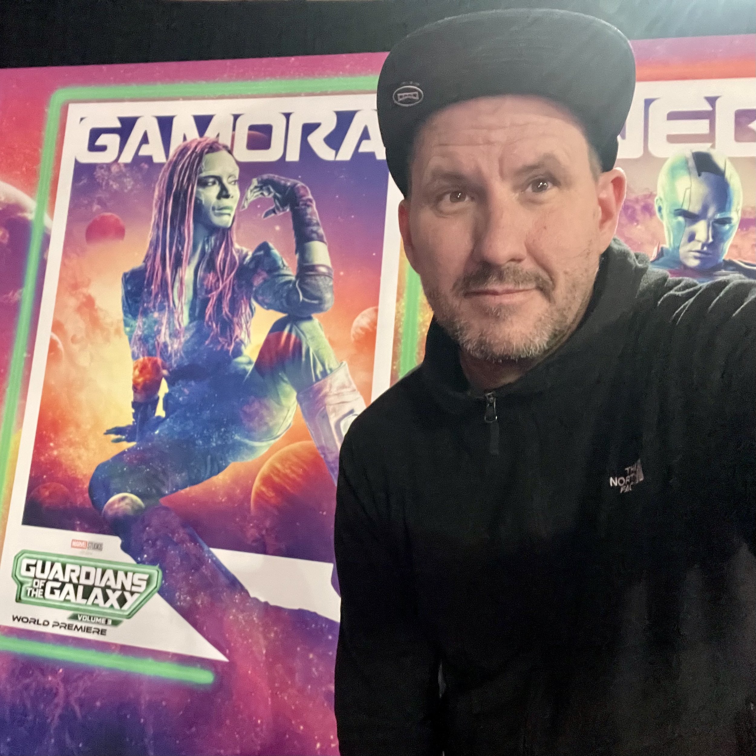 A man taking a selfie in front of a poster for the movie 'Guardians of the Galaxy', featuring a character with long pink hair and a cosmic background.