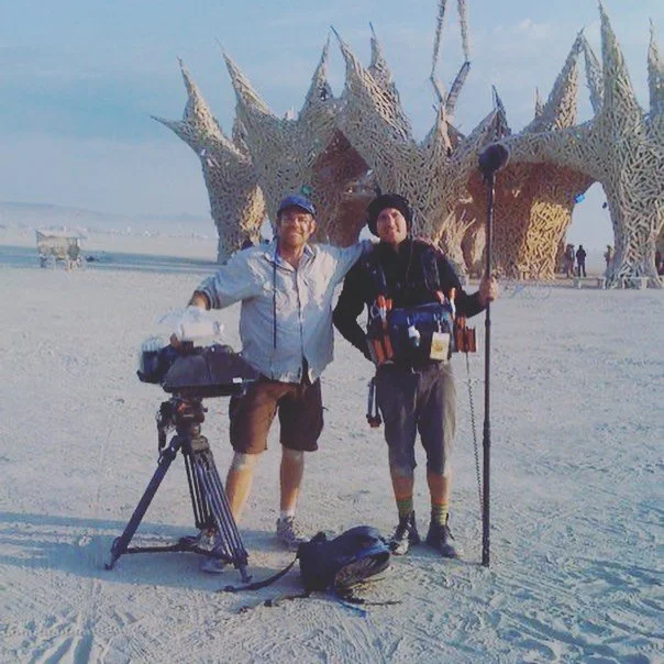 Two men standing in front of the Burning Man sculpture at Burning Man festival, one with a camera on a tripod and the other holding a walking stick, with a backpack and various gear.