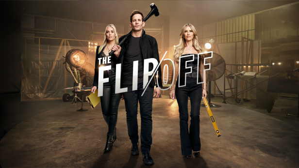 Three people standing in a studio with film equipment, holding signs that spell out "THE FLIP OFF," with bright studio lights and equipment in the background.