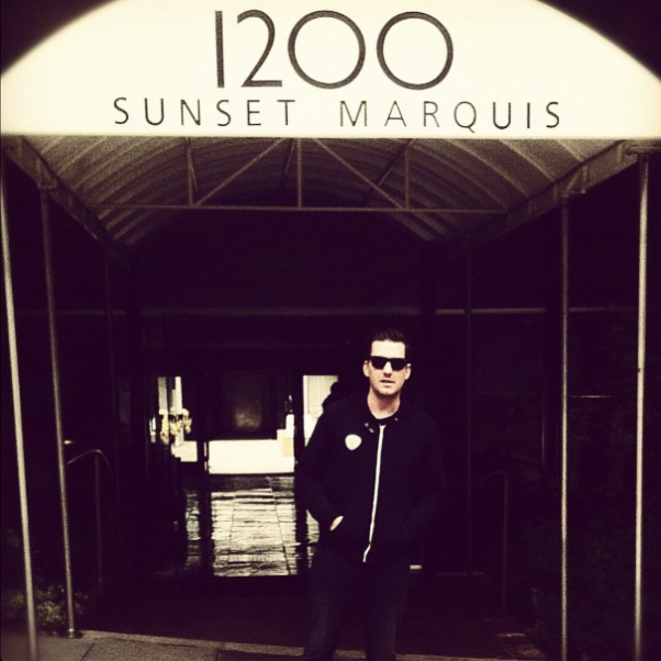 Person wearing black jacket and sunglasses standing under an archway with a sign that reads '1200 Sunset Marquis'.
