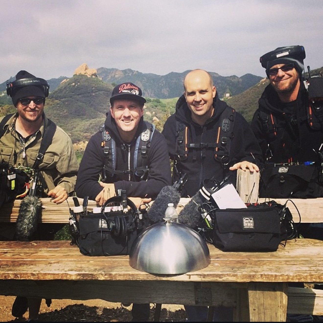 Four men with backpacks and outdoor gear smiling at a wooden table outdoors, with mountains in the background.