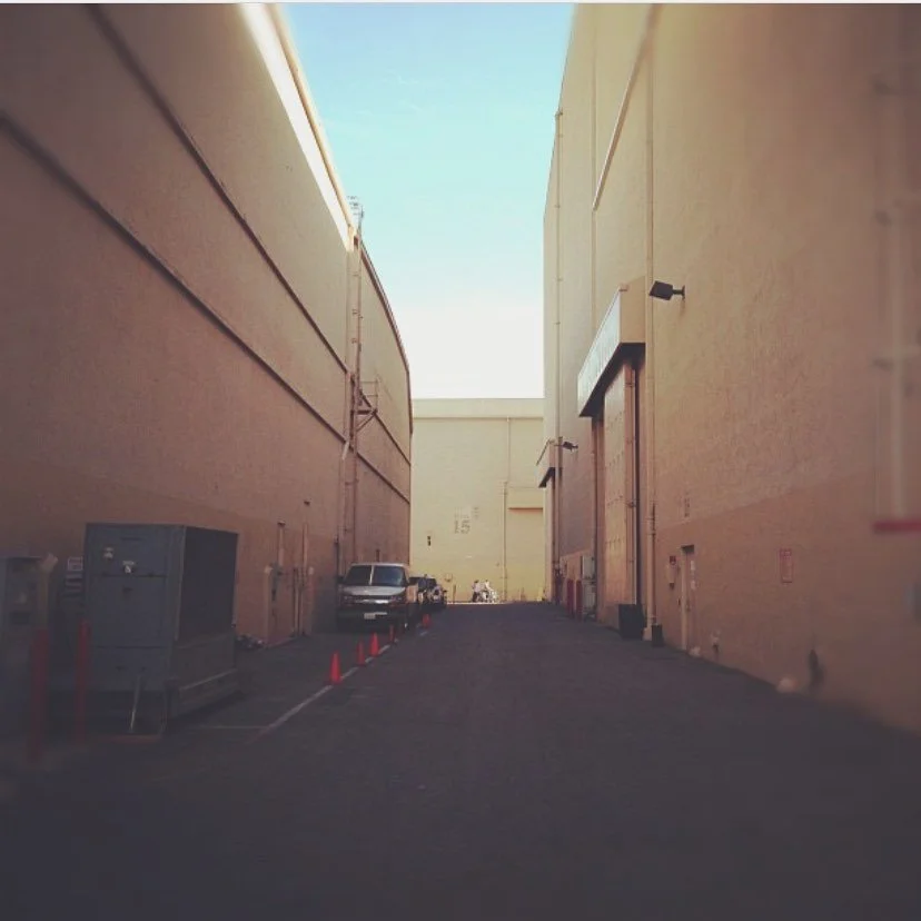 An alleyway between large beige industrial buildings with parked vehicles and orange traffic cones on the left side under a clear blue sky.