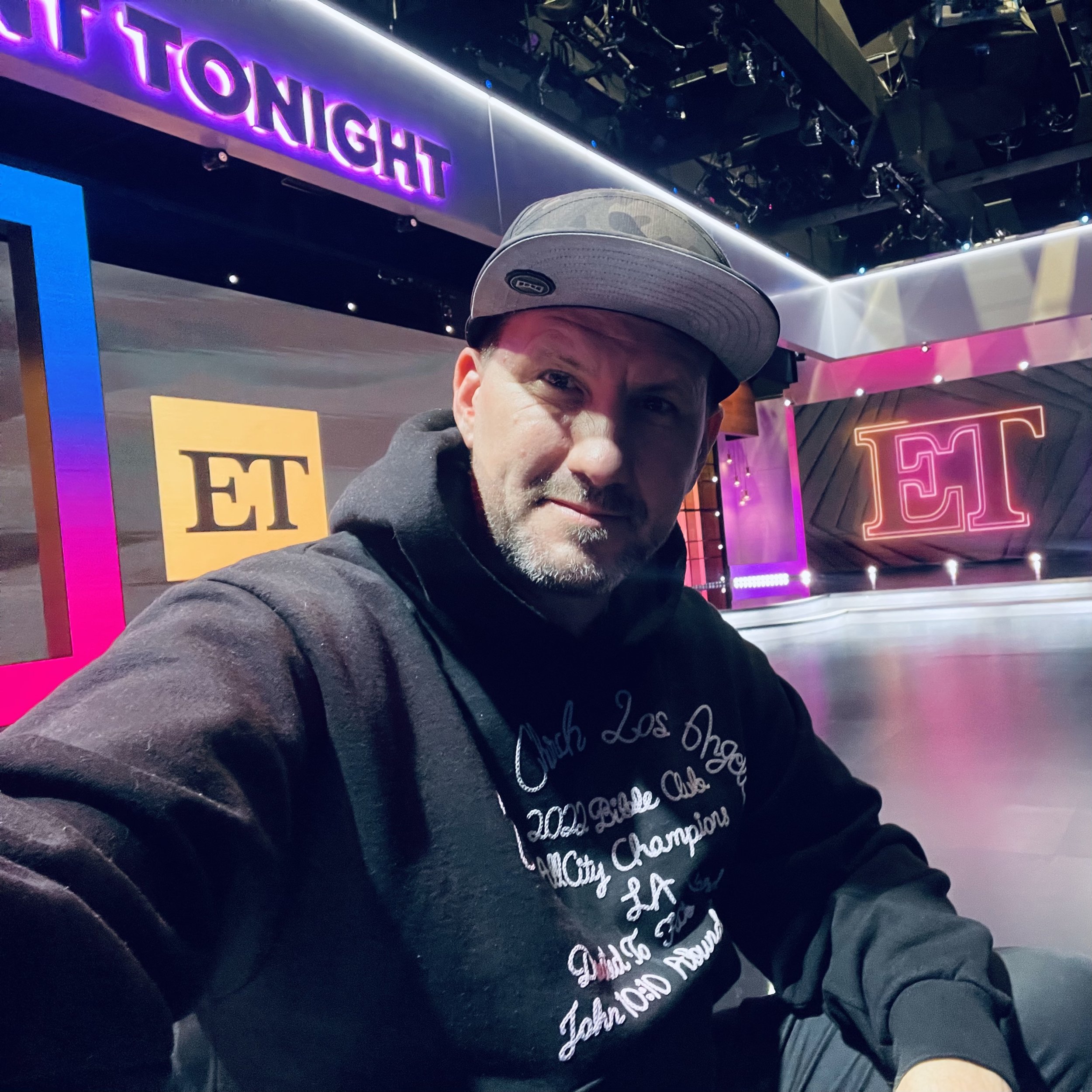 A man taking a selfie in a TV studio with ET signage and bright purple and pink lights in the background.