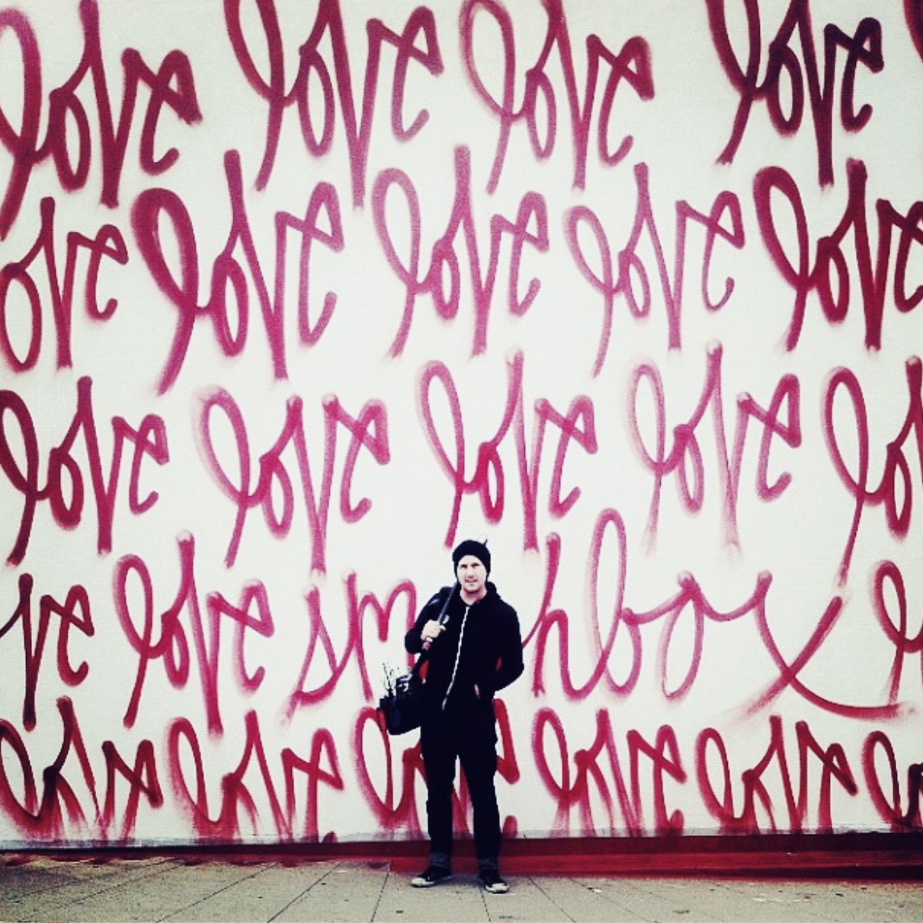 A person standing in front of a wall with repeated graffiti of the word "LOVE" in pink and purple spray paint.
