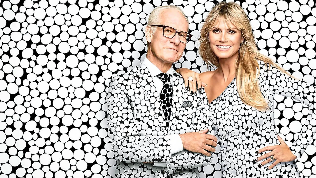 Two people, a man and a woman, posing together in front of a background covered with black and white polka dots. They are wearing matching polka dot outfits and smiling at the camera.