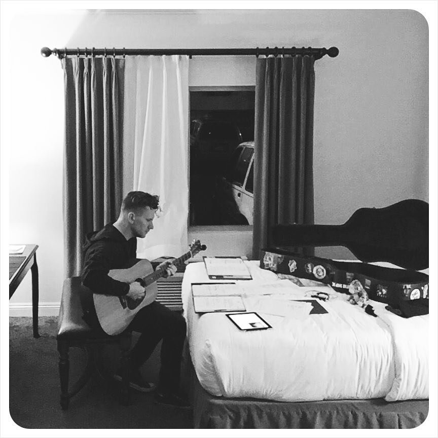 A man sitting on a chair playing an acoustic guitar in a hotel room with a window showing parked cars outside.