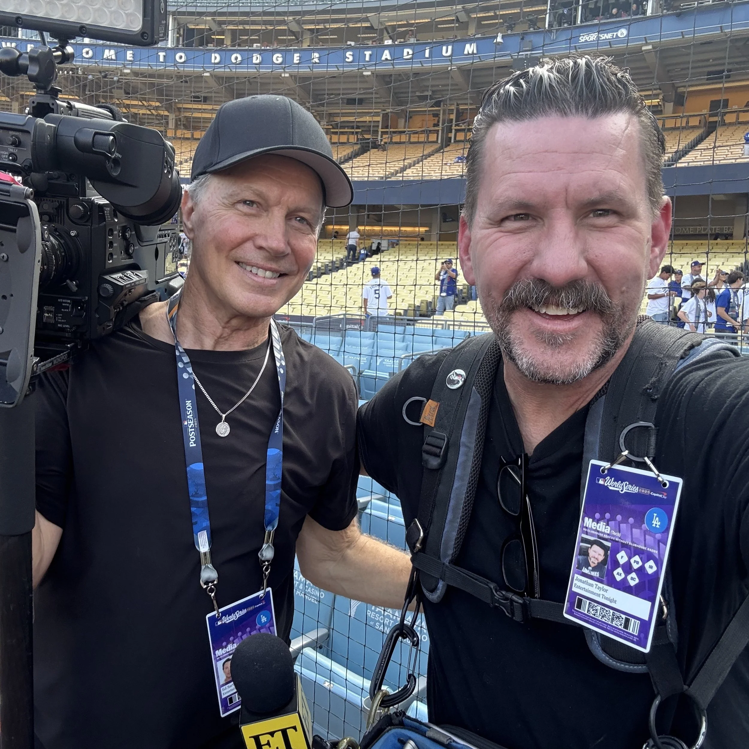 Two men taking a selfie at Dodger Stadium, with a baseball field and players in the background. One man is holding a camera and the other has a media pass hanging around his neck.