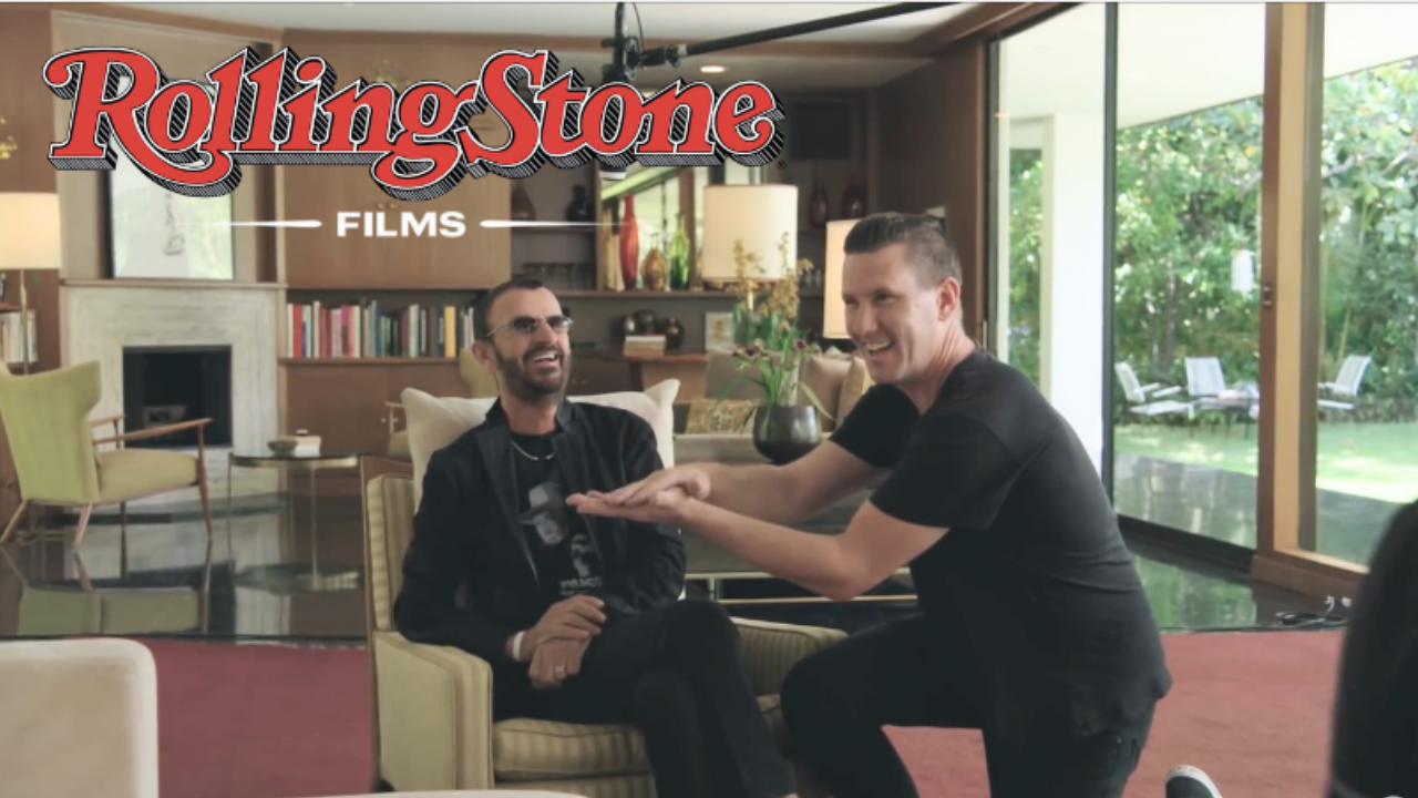 Two men laughing and playing rock-paper-scissors in a bright living room with large windows and outdoor patio in the background.