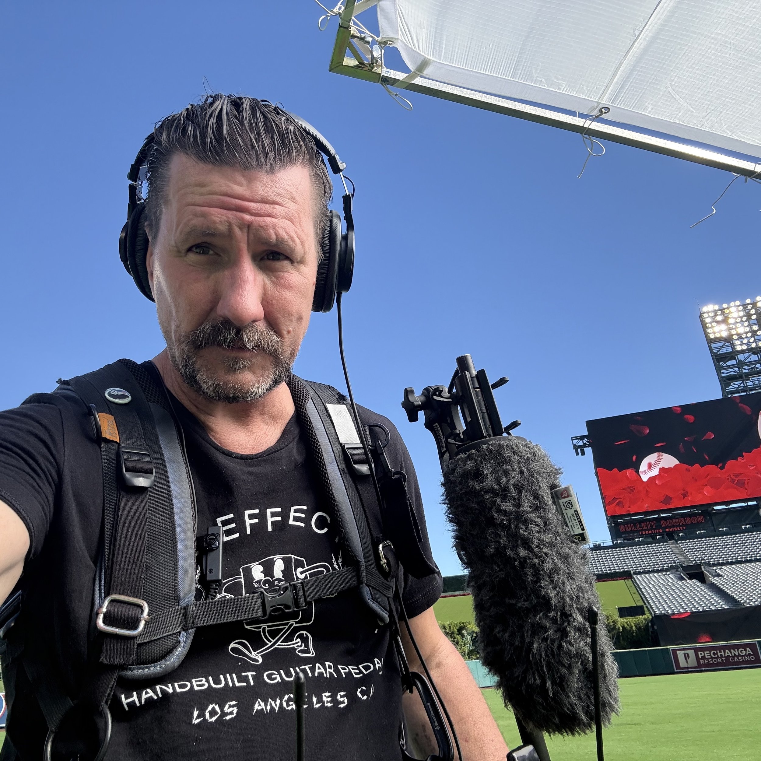 A man with a beard and headphones holding a microphone at a baseball field under a clear blue sky.