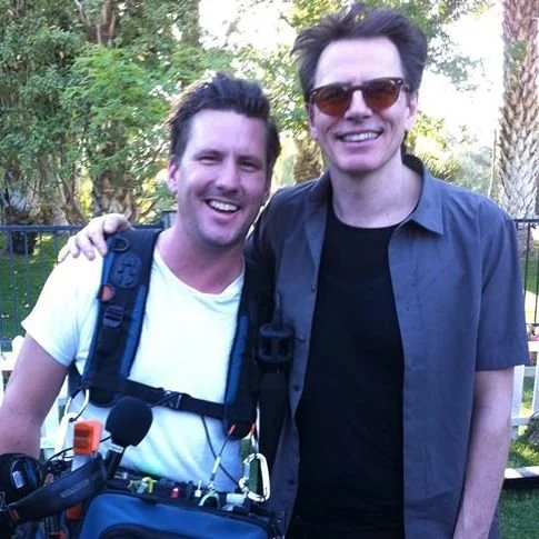 Two men smiling outdoors, one with a backpack and equipment, and the other wearing sunglasses, standing close together.
