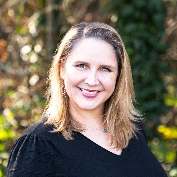 A smiling woman with shoulder-length blonde hair, wearing a black top, standing outdoors with a blurred background of trees and sunlight.