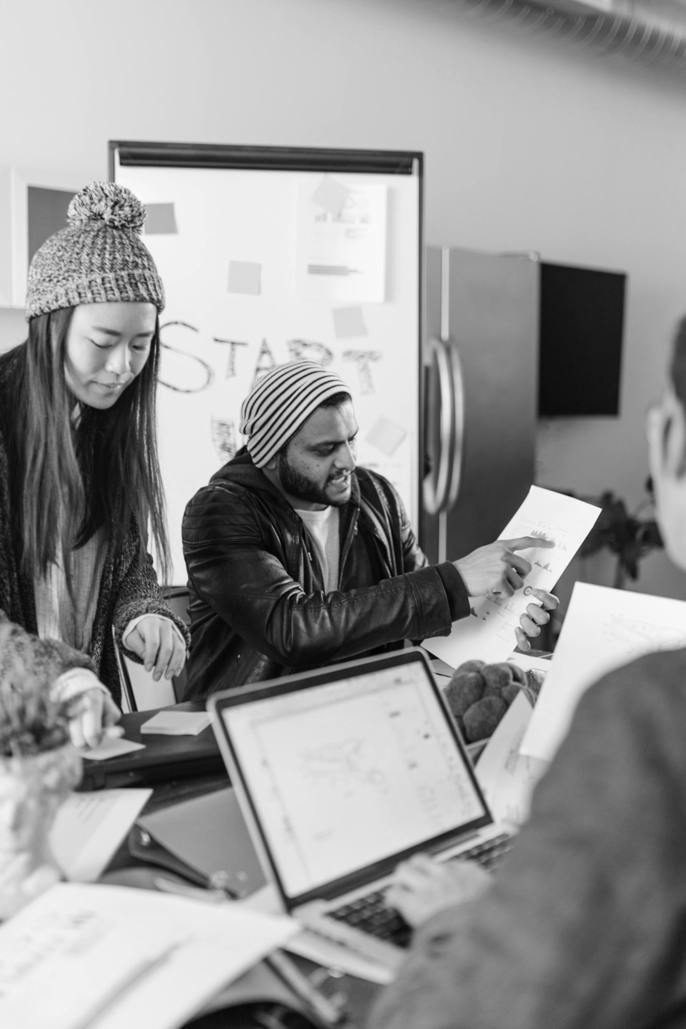 Black and white photo of a group of young adults collaborating in an office, wearing winter hats, with a whiteboard in the background and laptops on the table.