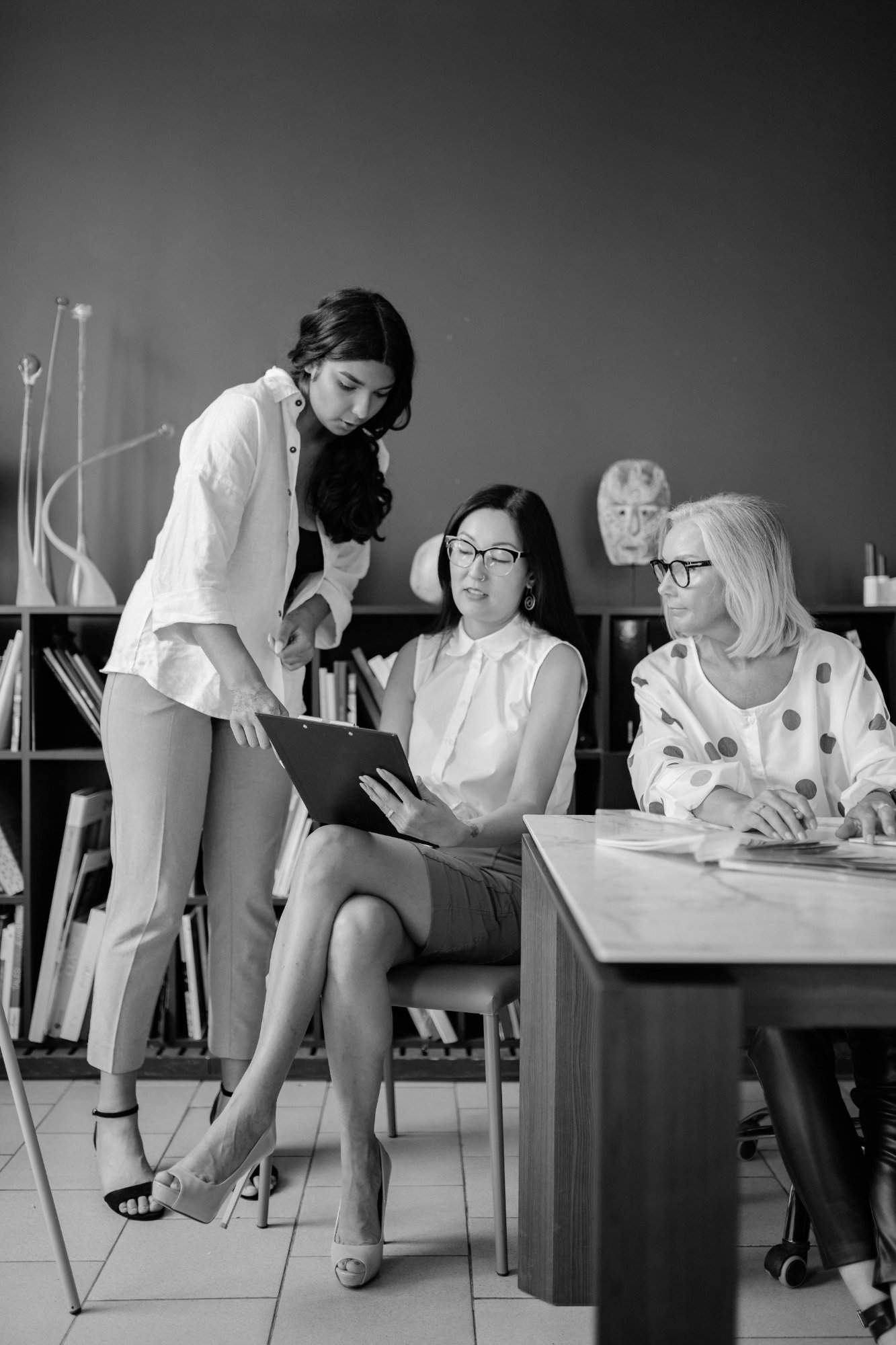 Three women in an office engaged in a discussion. One woman is standing and pointing to a tablet, while the other two women are seated, looking at the tablet.