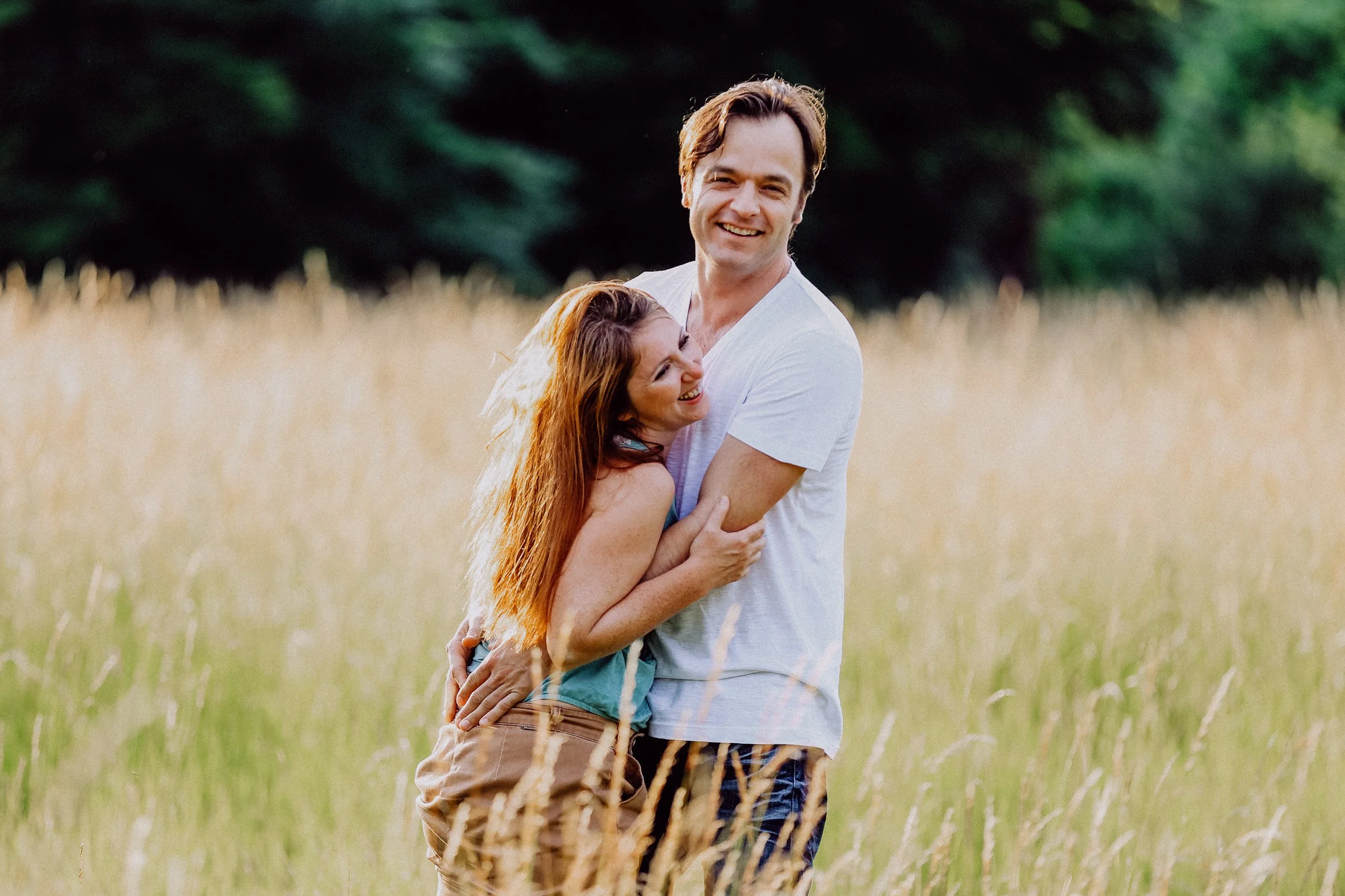 Ein lachendes Paar steht in einem Feld mit hohen Gräsern, im Hintergrund Bäume, bei Sonnenuntergang.